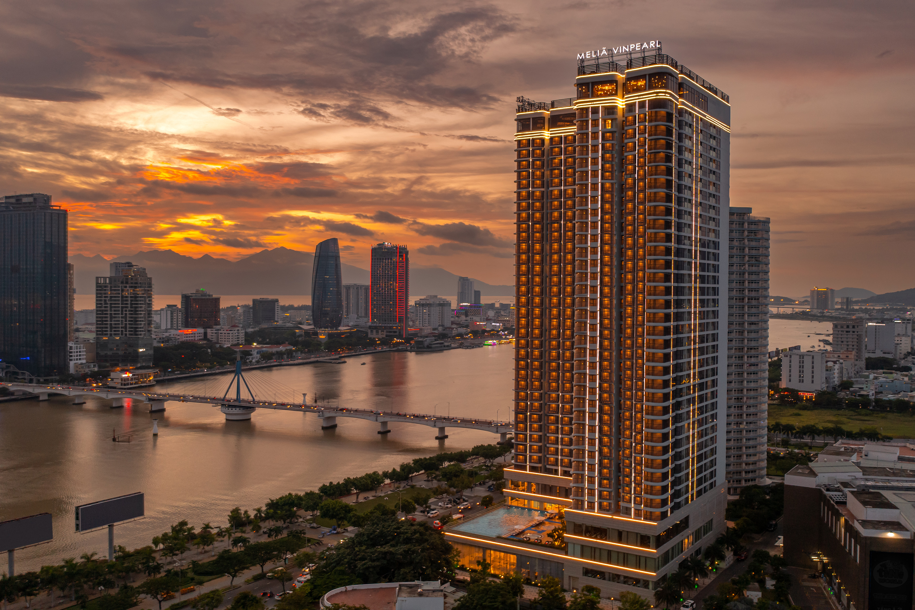 Da Nang Meliá Vinpearl hotel by river at sunset, city lights, mountains.