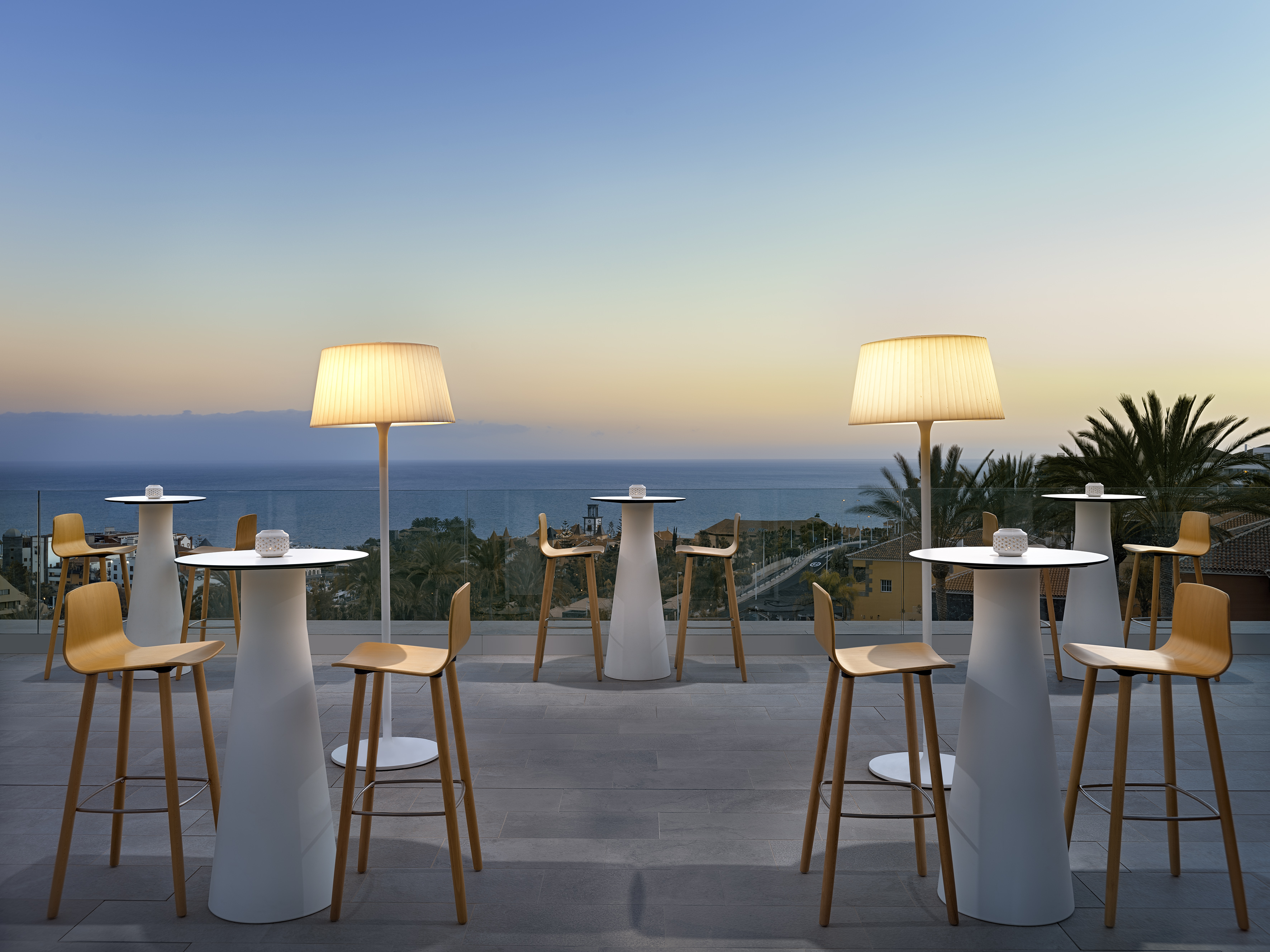 Rooftop terrace bar with sunset ocean view, high tables, and floor lamps.