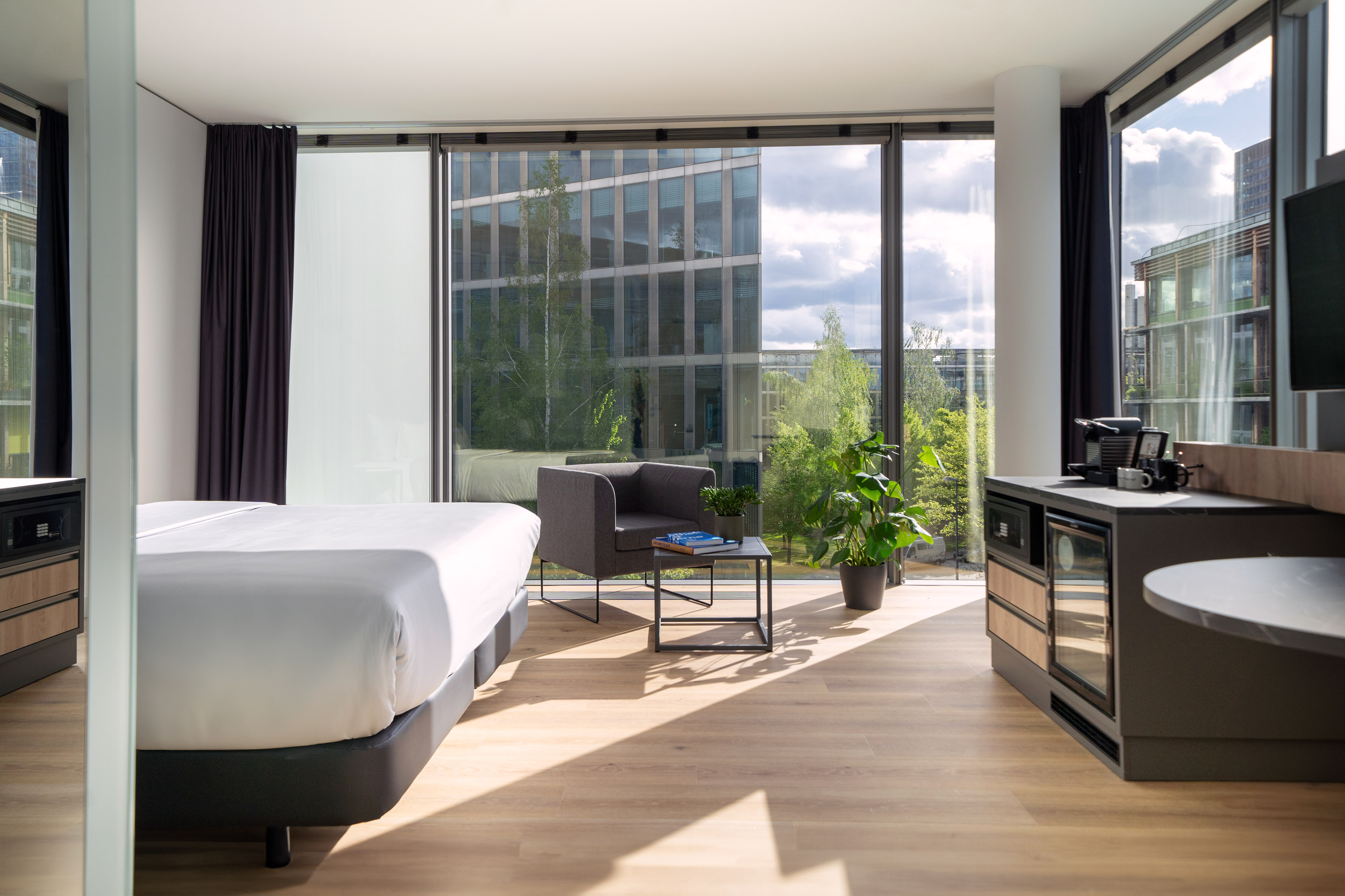 Modern hotel room with bed, armchair, wood floor, and bright city views.