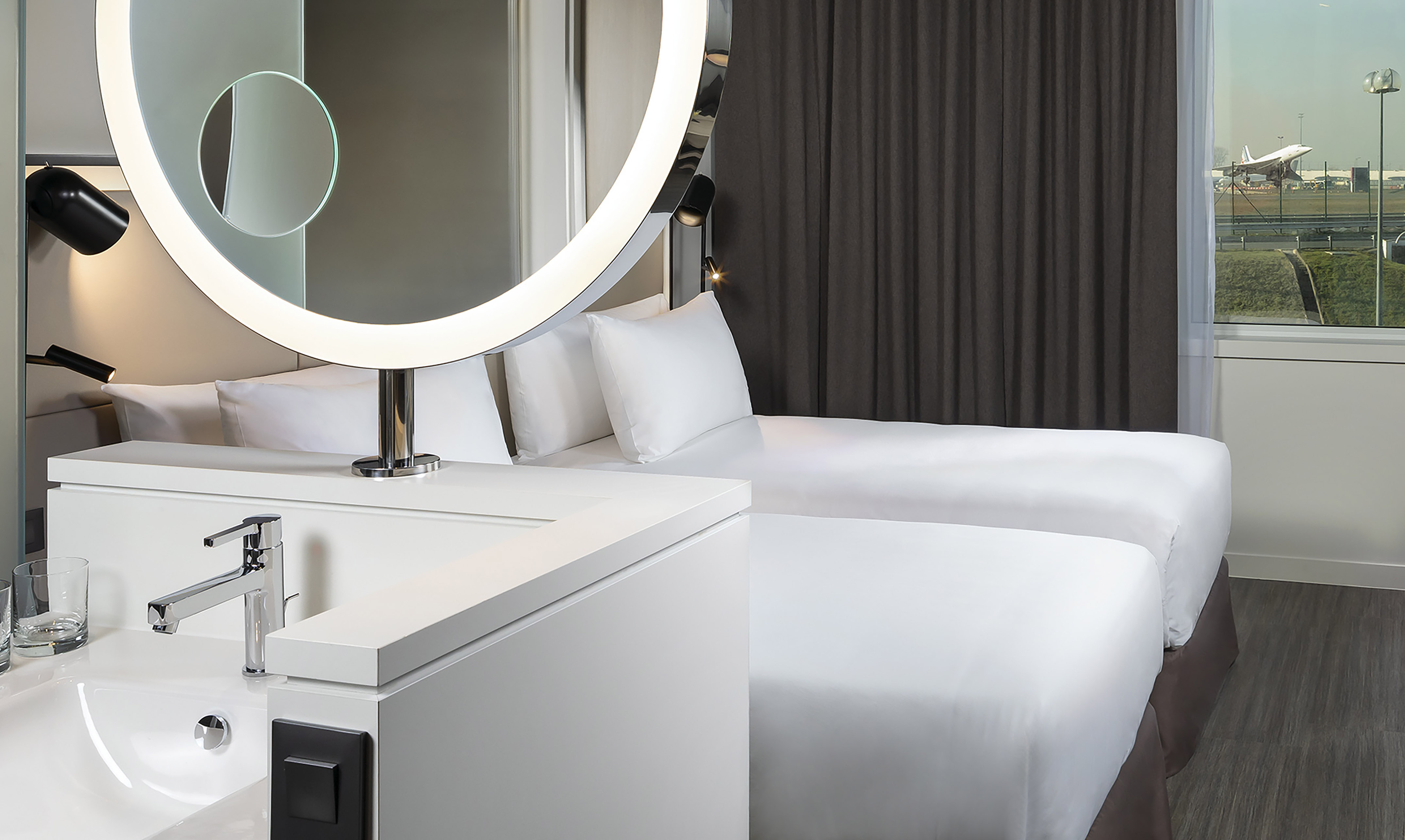 Modern hotel room: twin beds, illuminated mirror, airport view with plane.