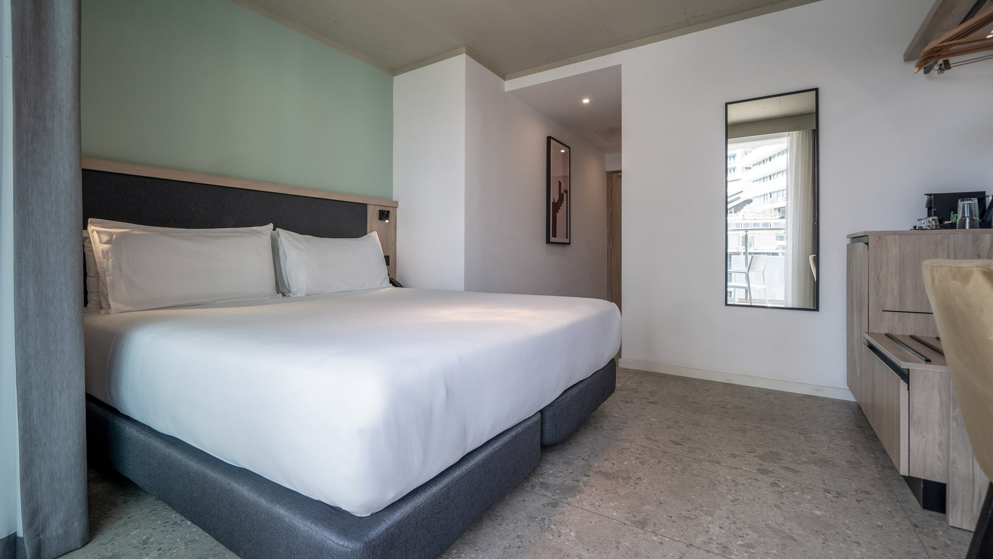 Modern hotel room with large bed, white bedding, green accent wall, grey floor.