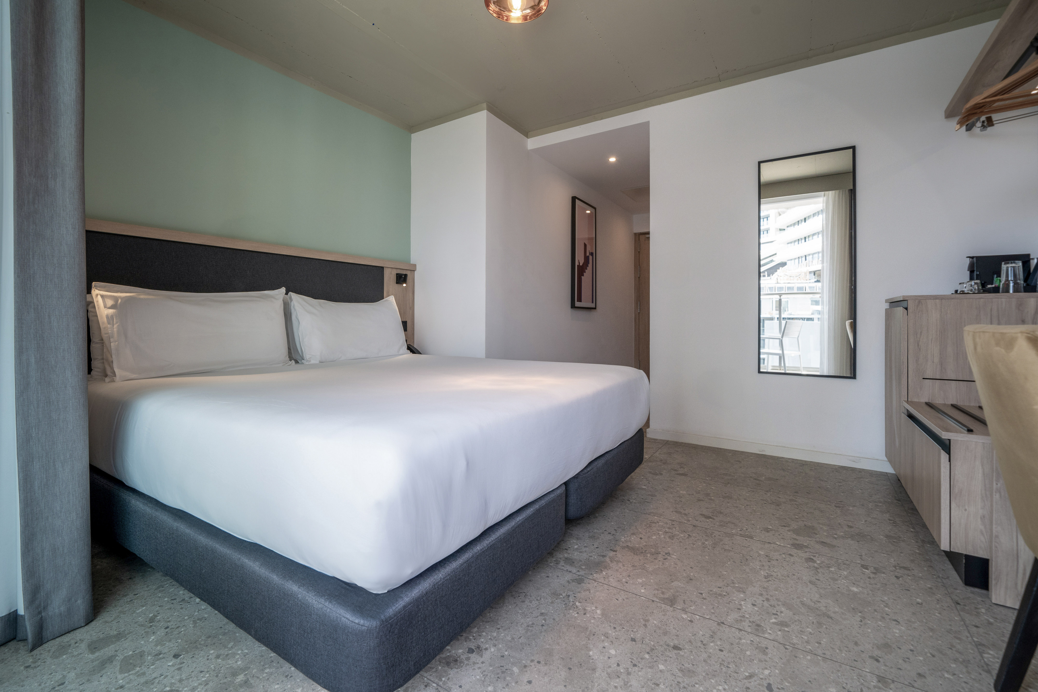 Modern hotel room with large bed, white bedding, green accent wall, grey floor.