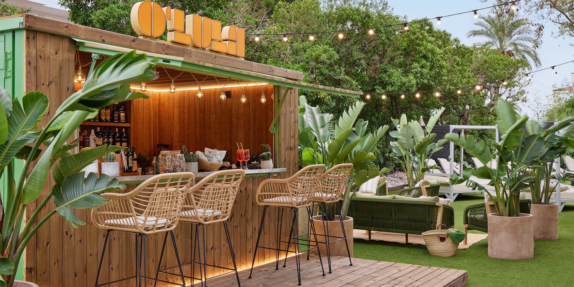 Tropical resort bar with wooden counter, wicker stools, and lush plants.