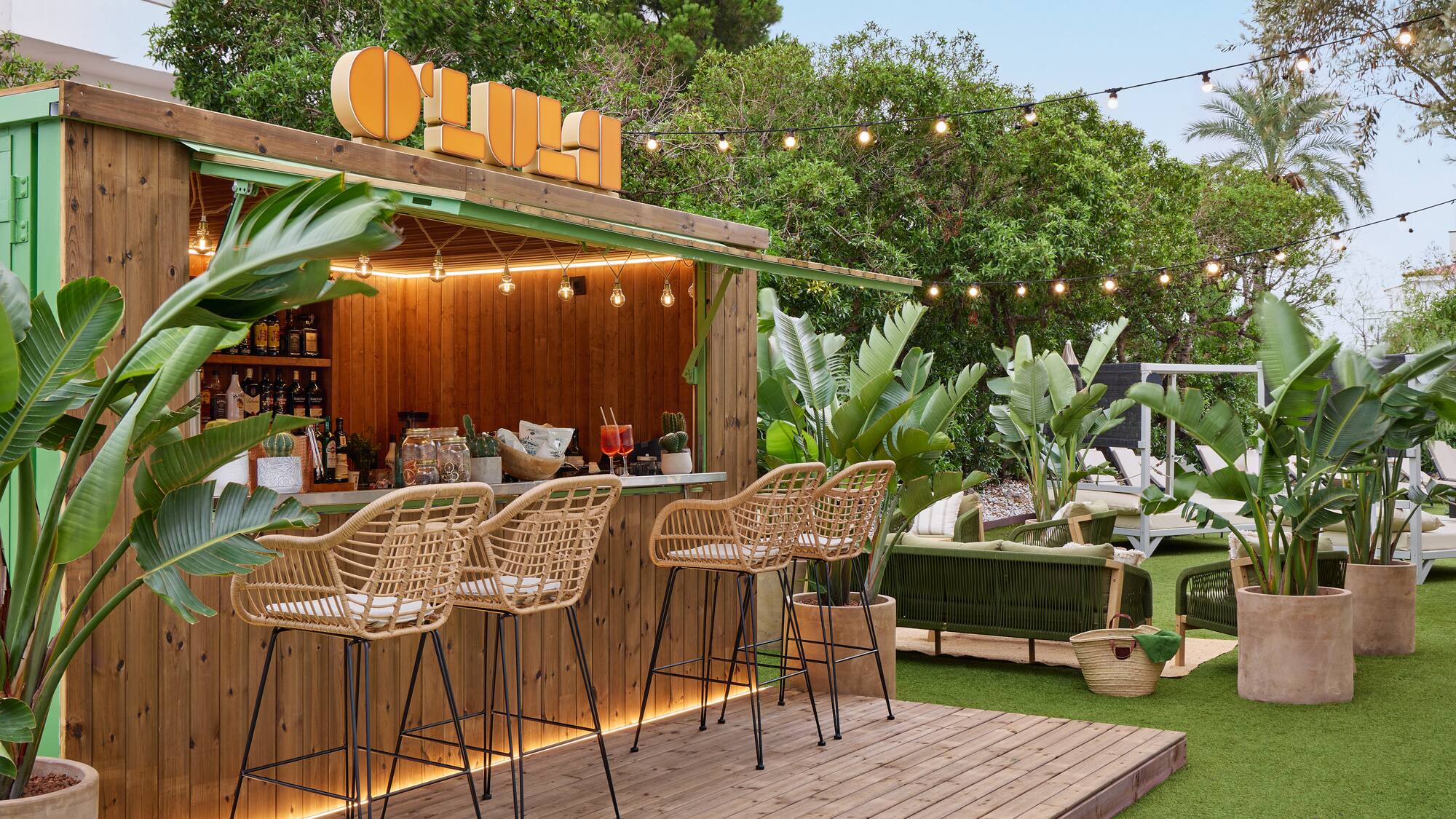 Tropical resort bar with wooden counter, wicker stools, and lush plants.