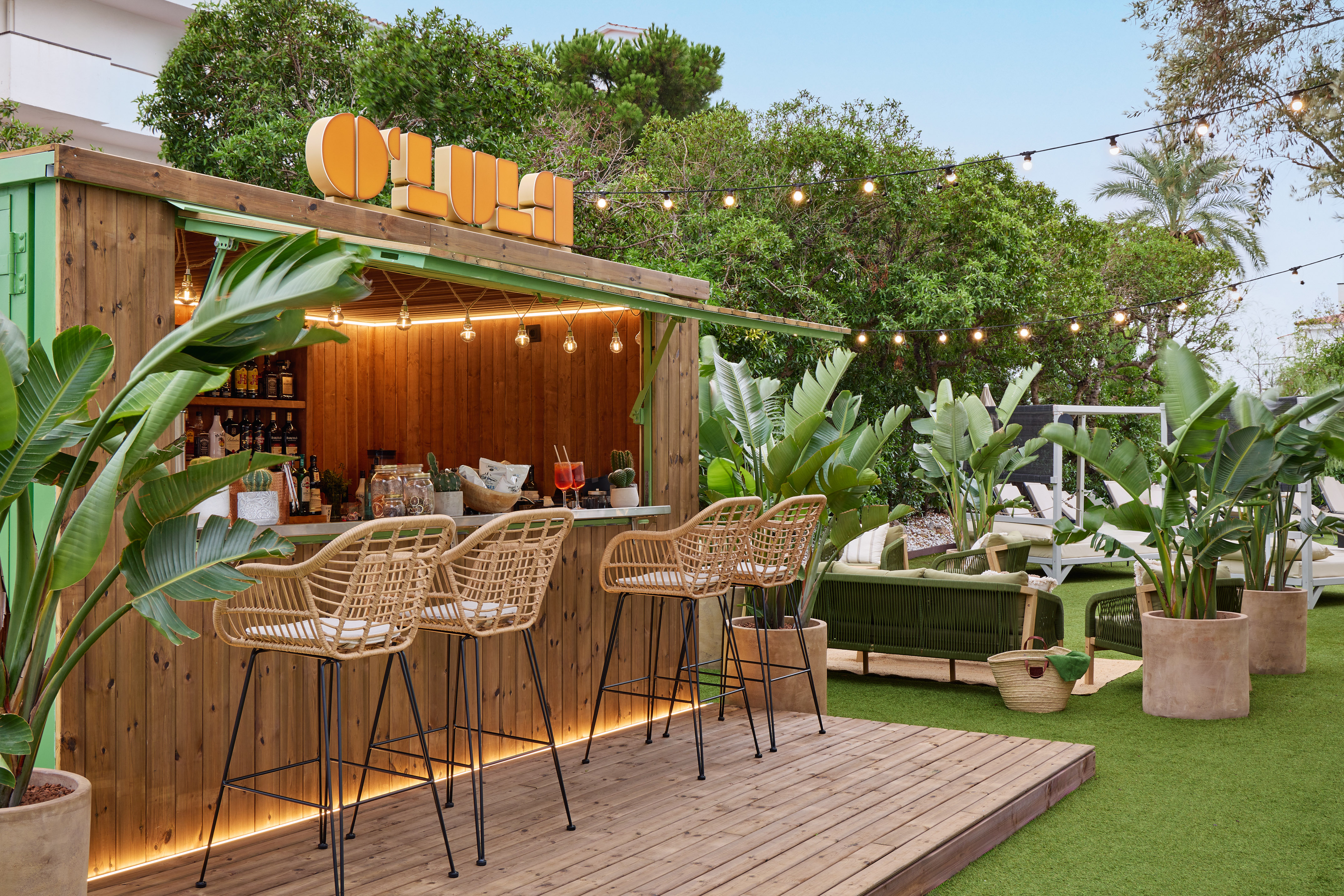 Tropical resort bar with wooden counter, wicker stools, and lush plants.