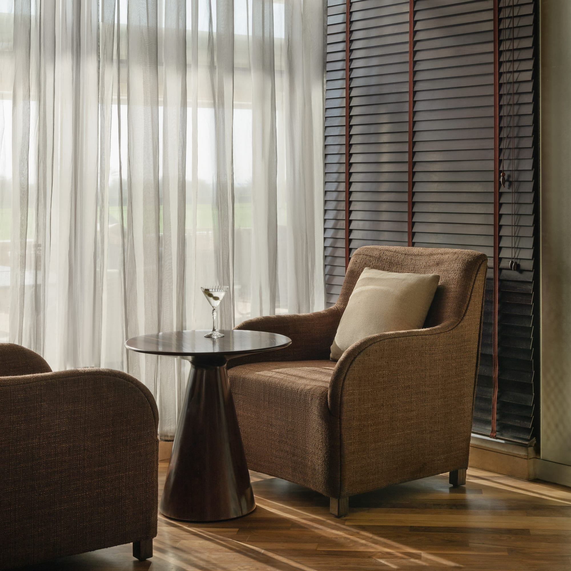 Lounge with brown armchairs, side table, martini, sheer curtains, and dark blinds.