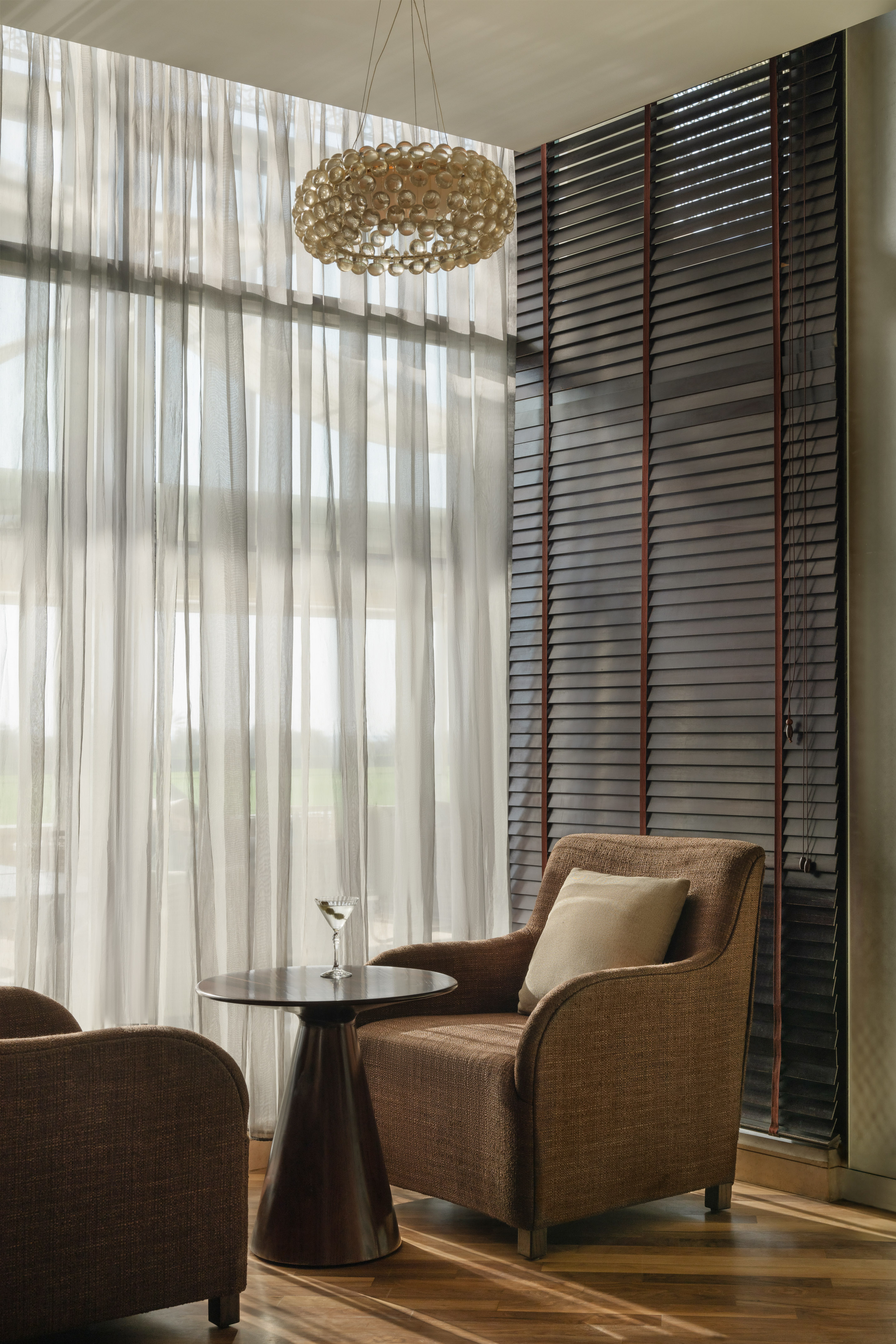 Lounge with brown armchairs, side table, martini, sheer curtains, and dark blinds.