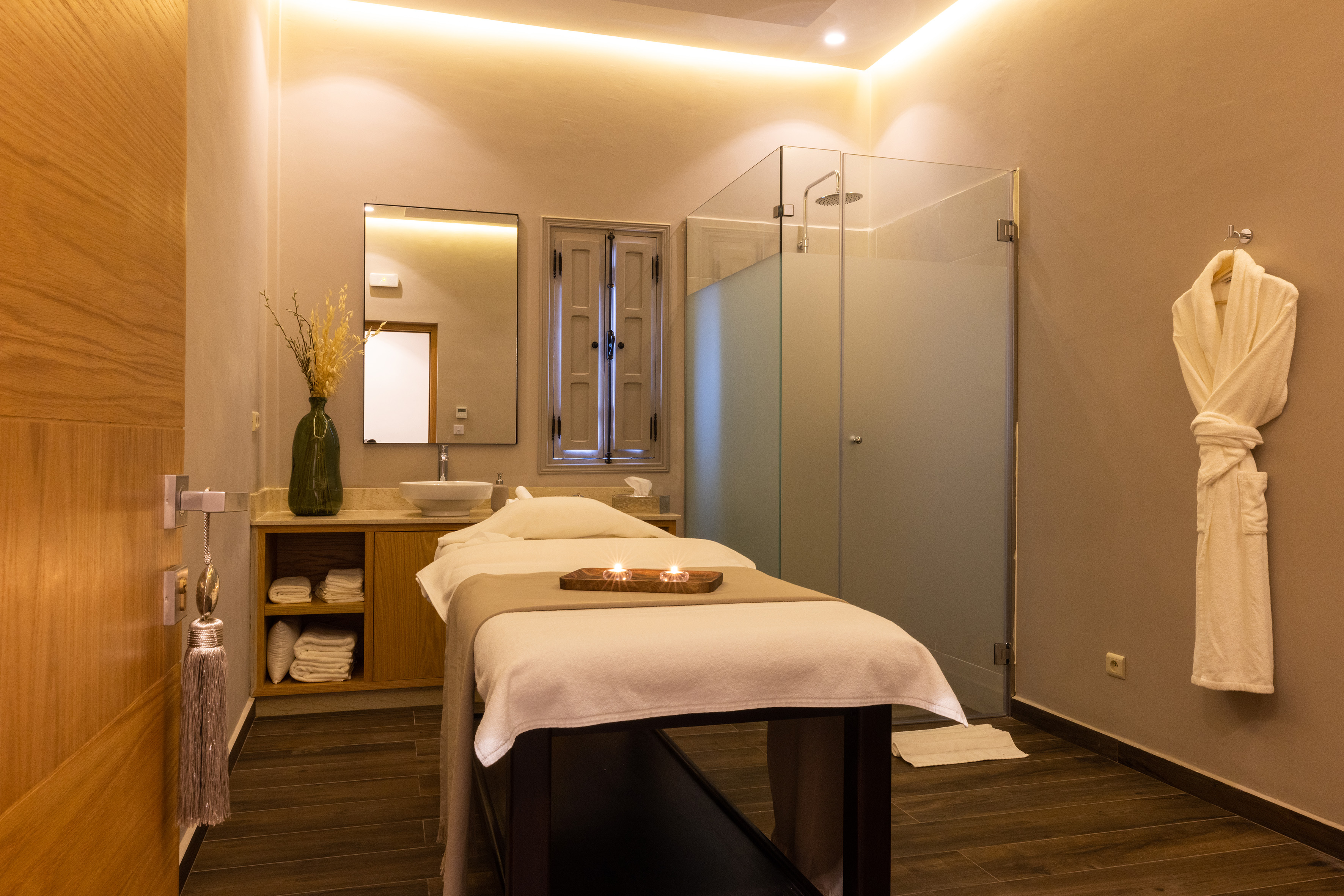 Spa treatment room with massage bed, glass shower, sink, and glowing candles.