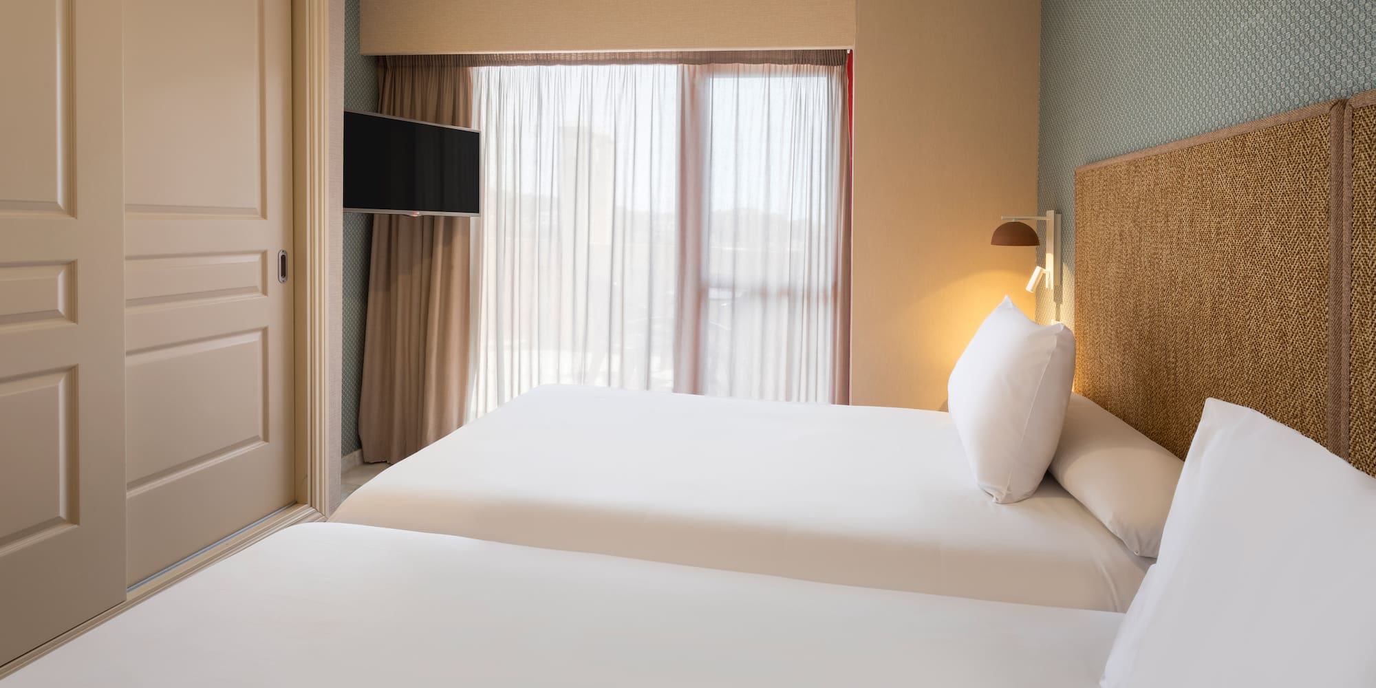 Hotel bedroom with twin beds, white linen, woven headboard, and wall TV.