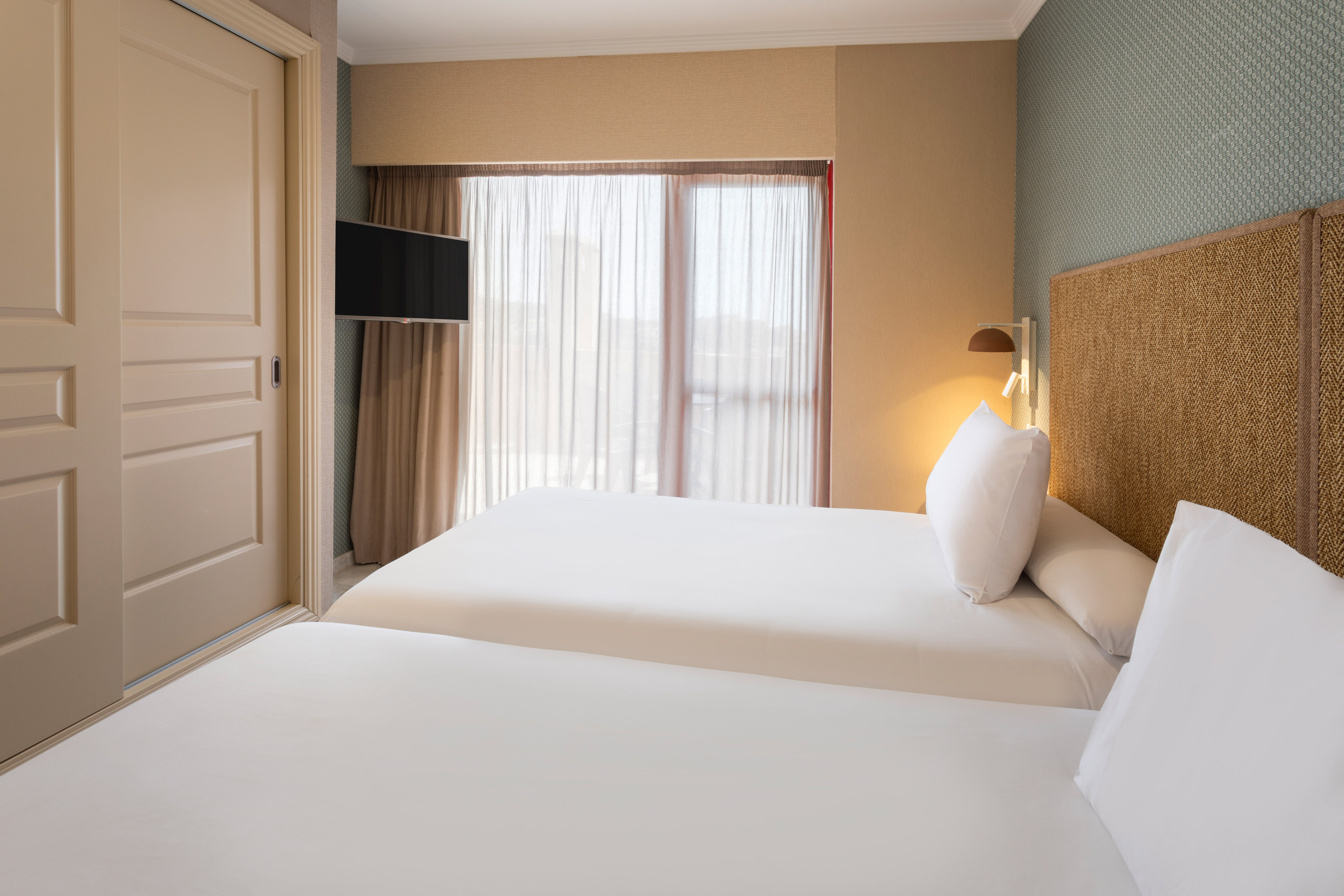 Hotel bedroom with twin beds, white linen, woven headboard, and wall TV.