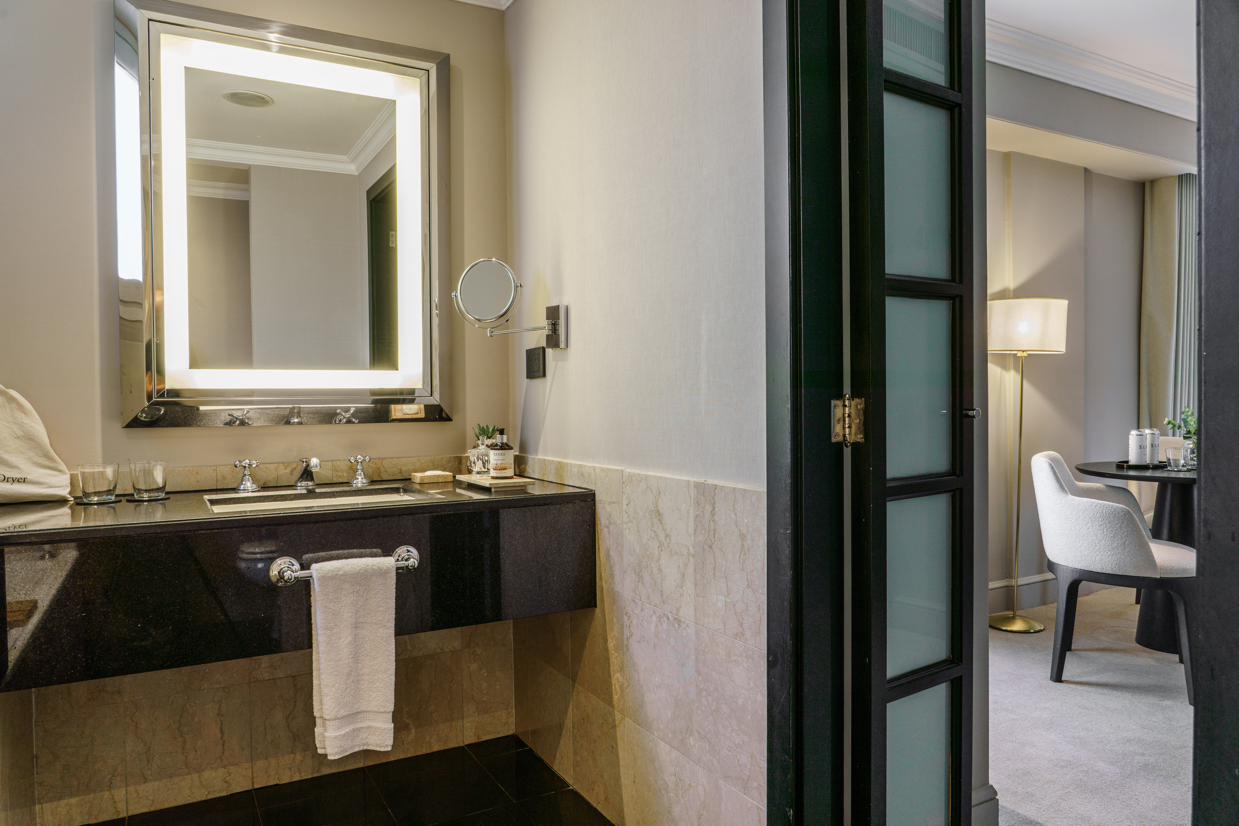 Modern hotel bathroom with lighted mirror, black vanity, and adjacent suite.