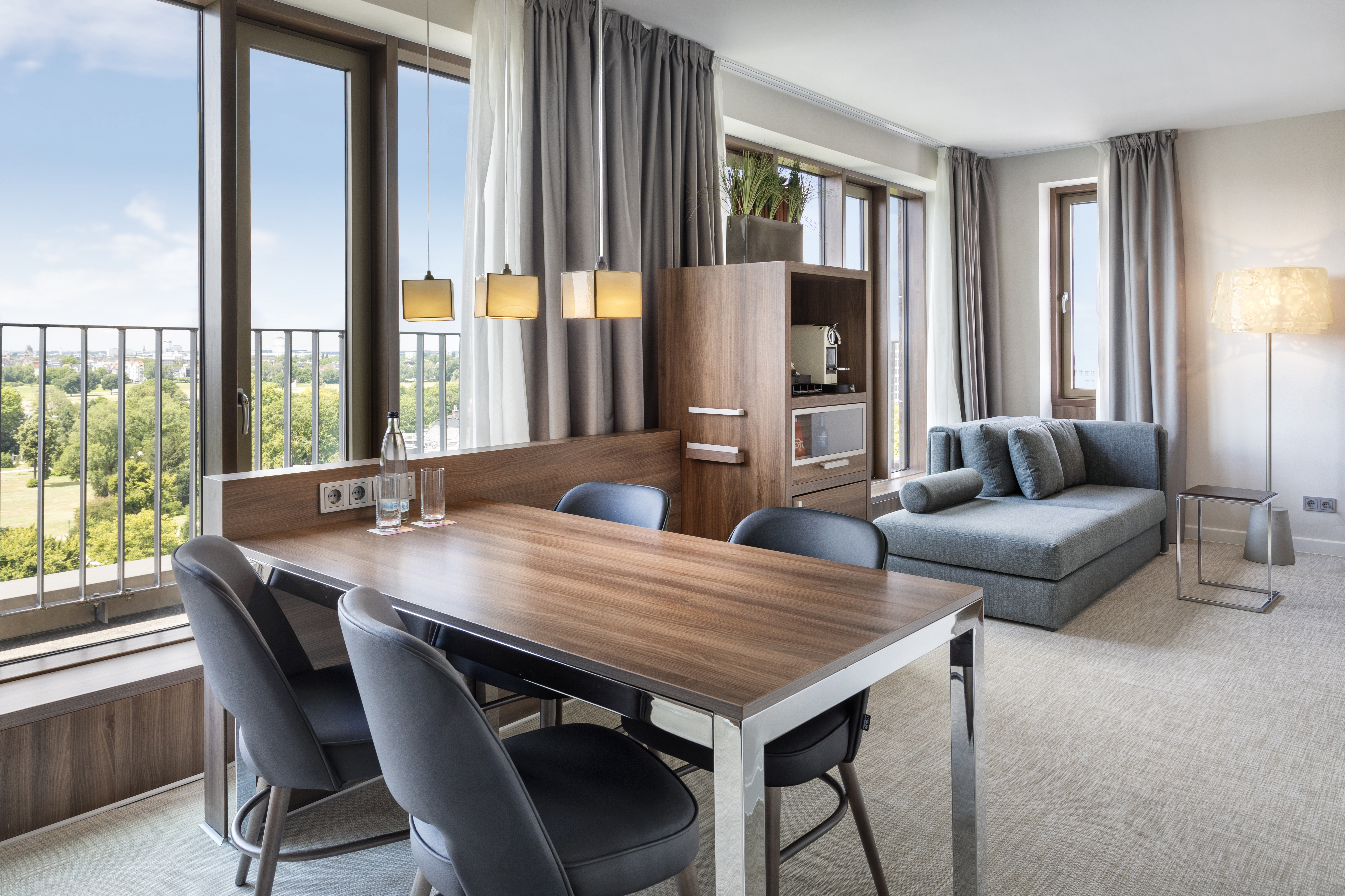 Modern hotel suite with dining table, sofa lounge, and green park view.