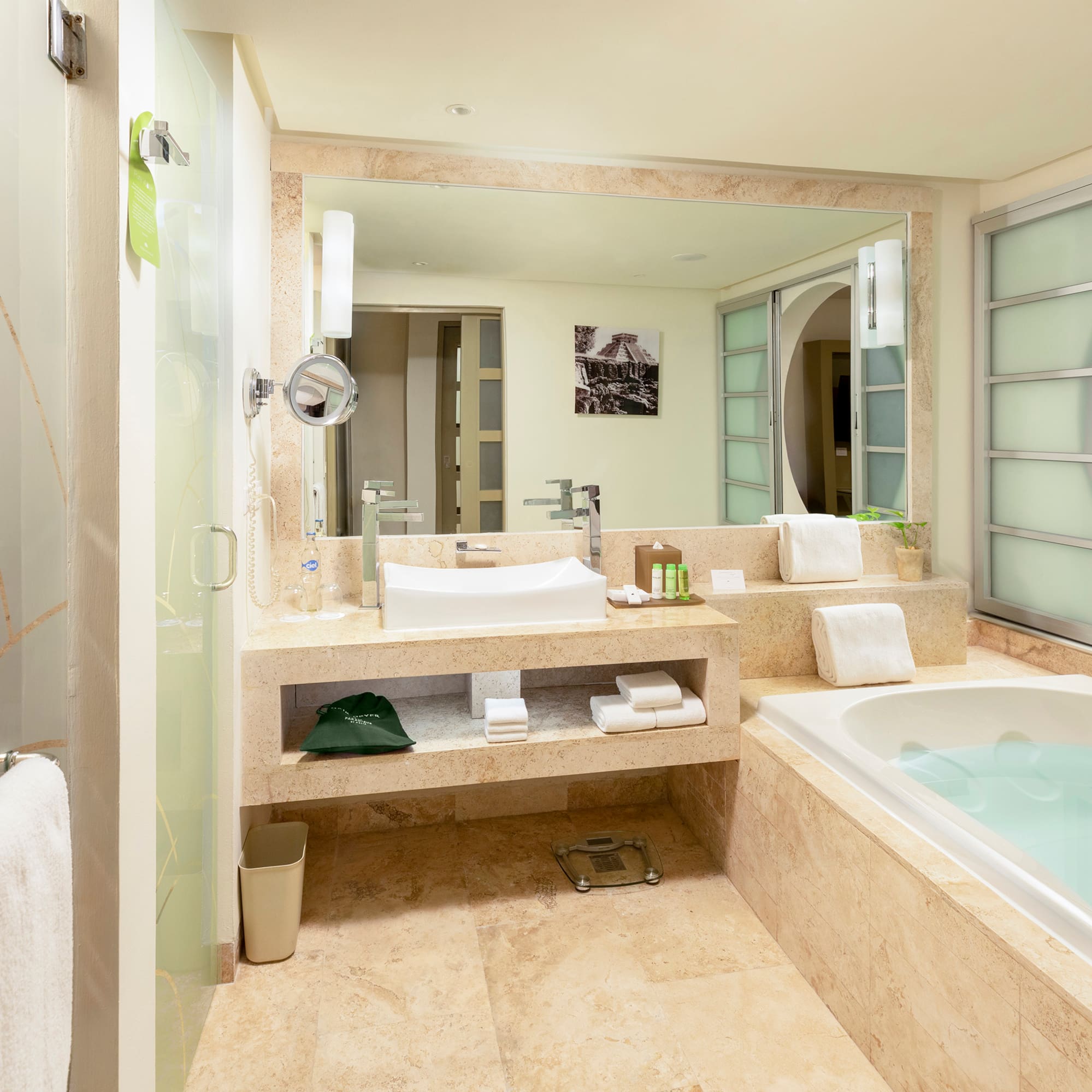 Modern hotel bathroom with tub, sink, glass shower, and bedroom view.