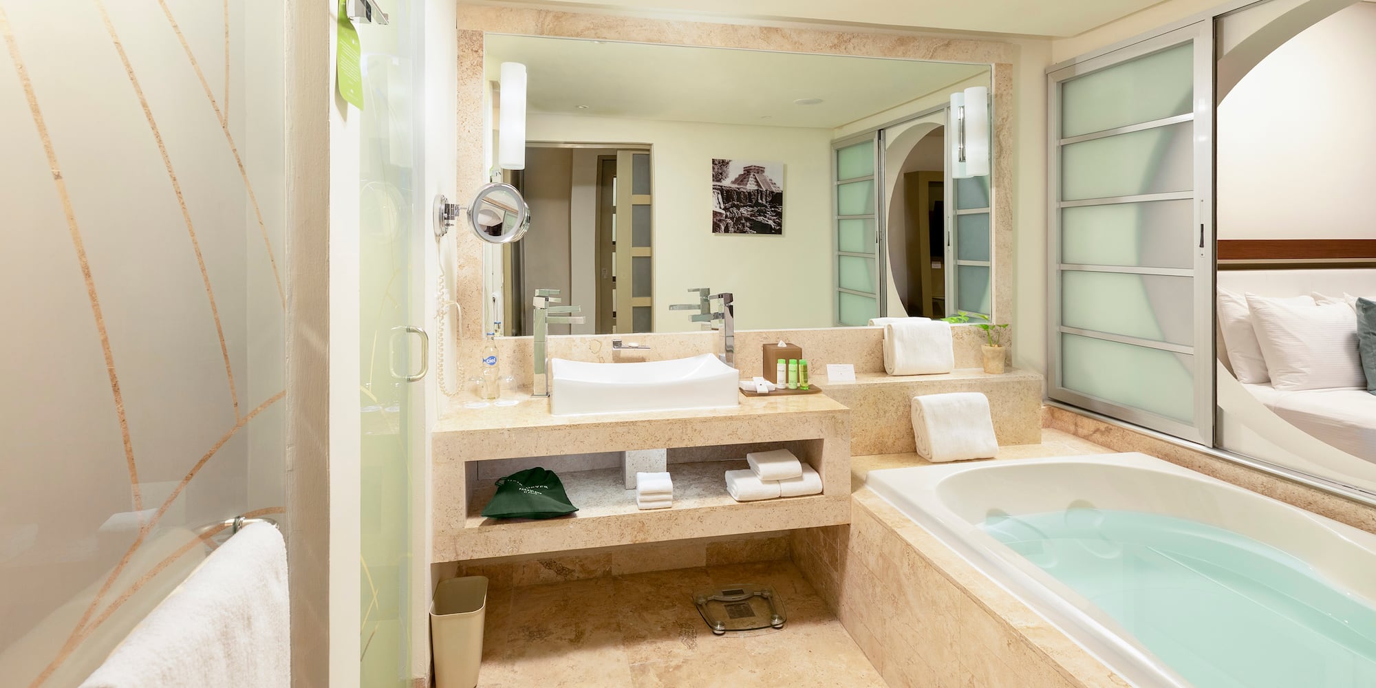 Modern hotel bathroom with tub, sink, glass shower, and bedroom view.