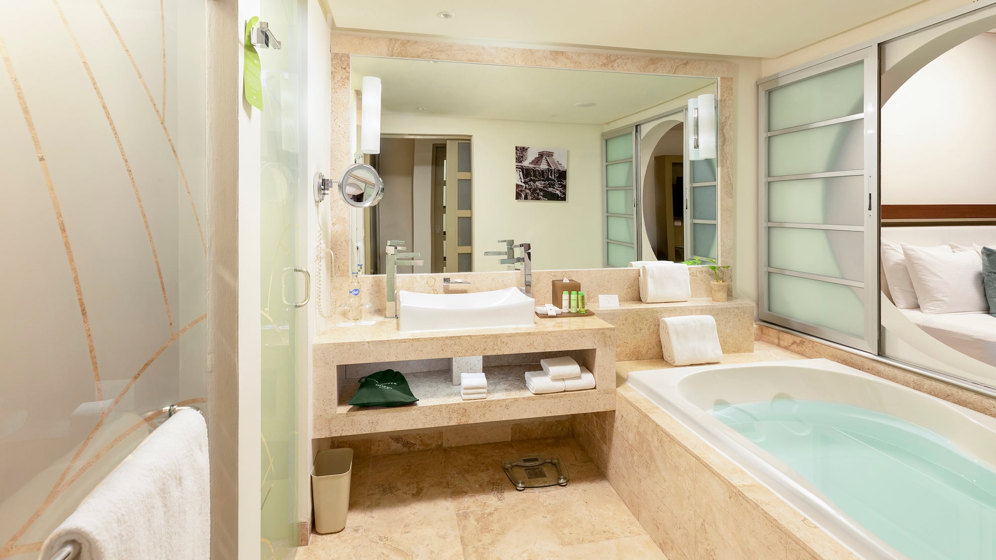 Modern hotel bathroom with tub, sink, glass shower, and bedroom view.