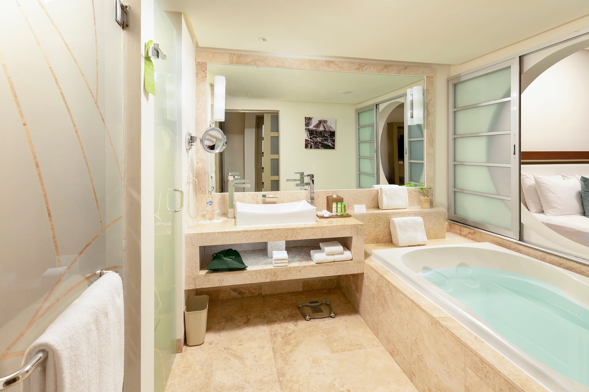 Modern hotel bathroom with tub, sink, glass shower, and bedroom view.