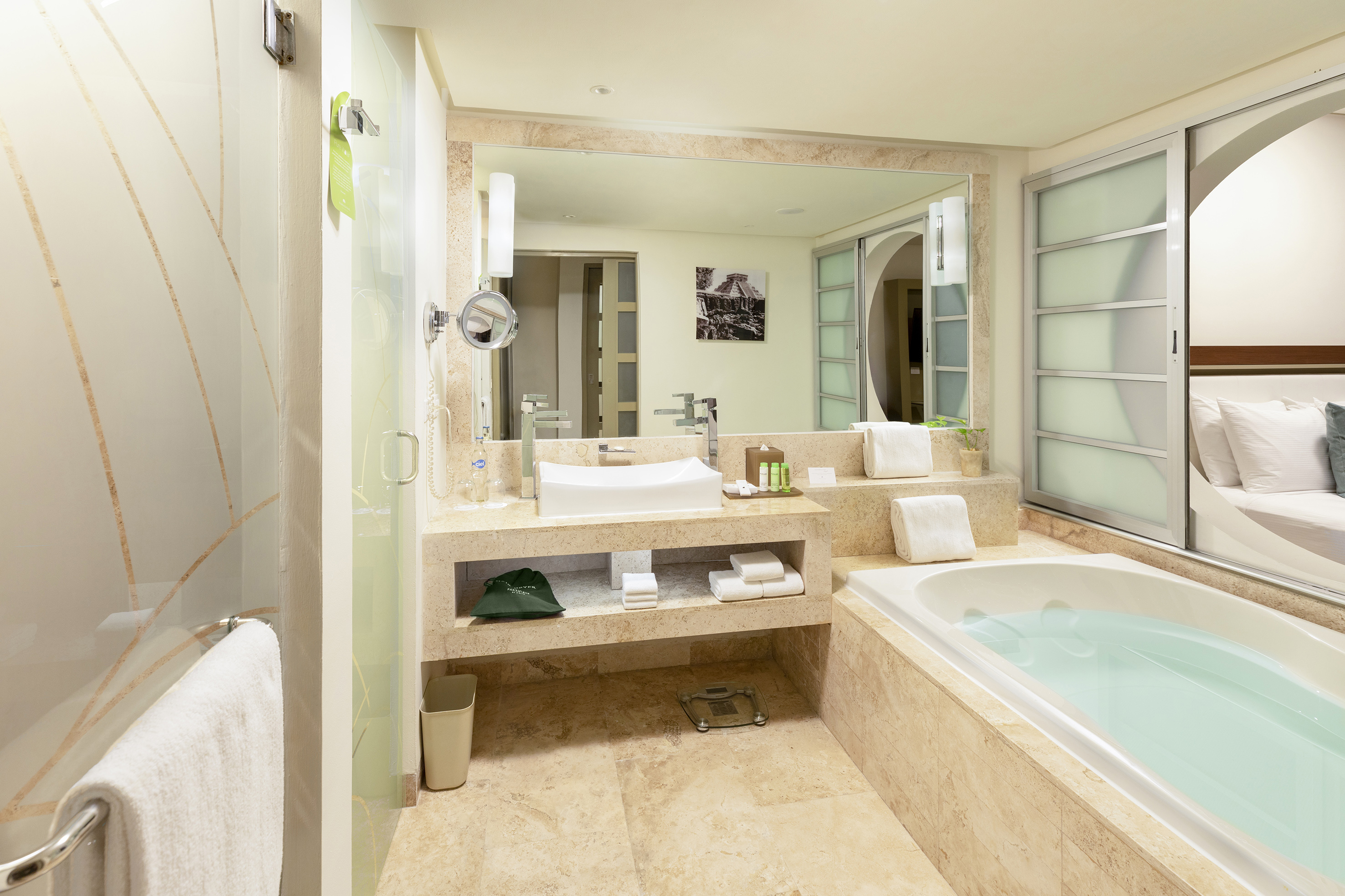 Modern hotel bathroom with tub, sink, glass shower, and bedroom view.
