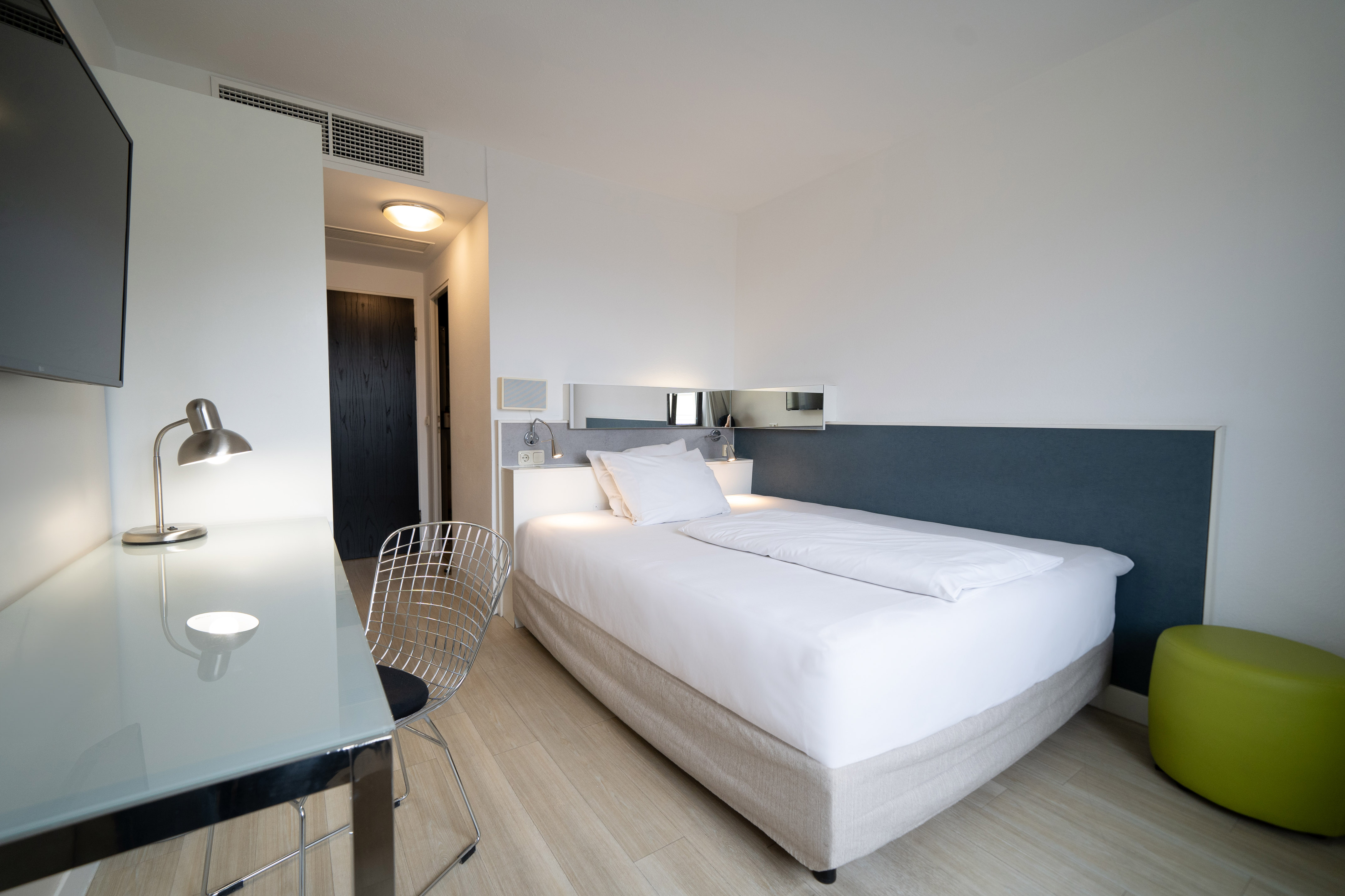 Modern single hotel room with desk, TV, and green ottoman.