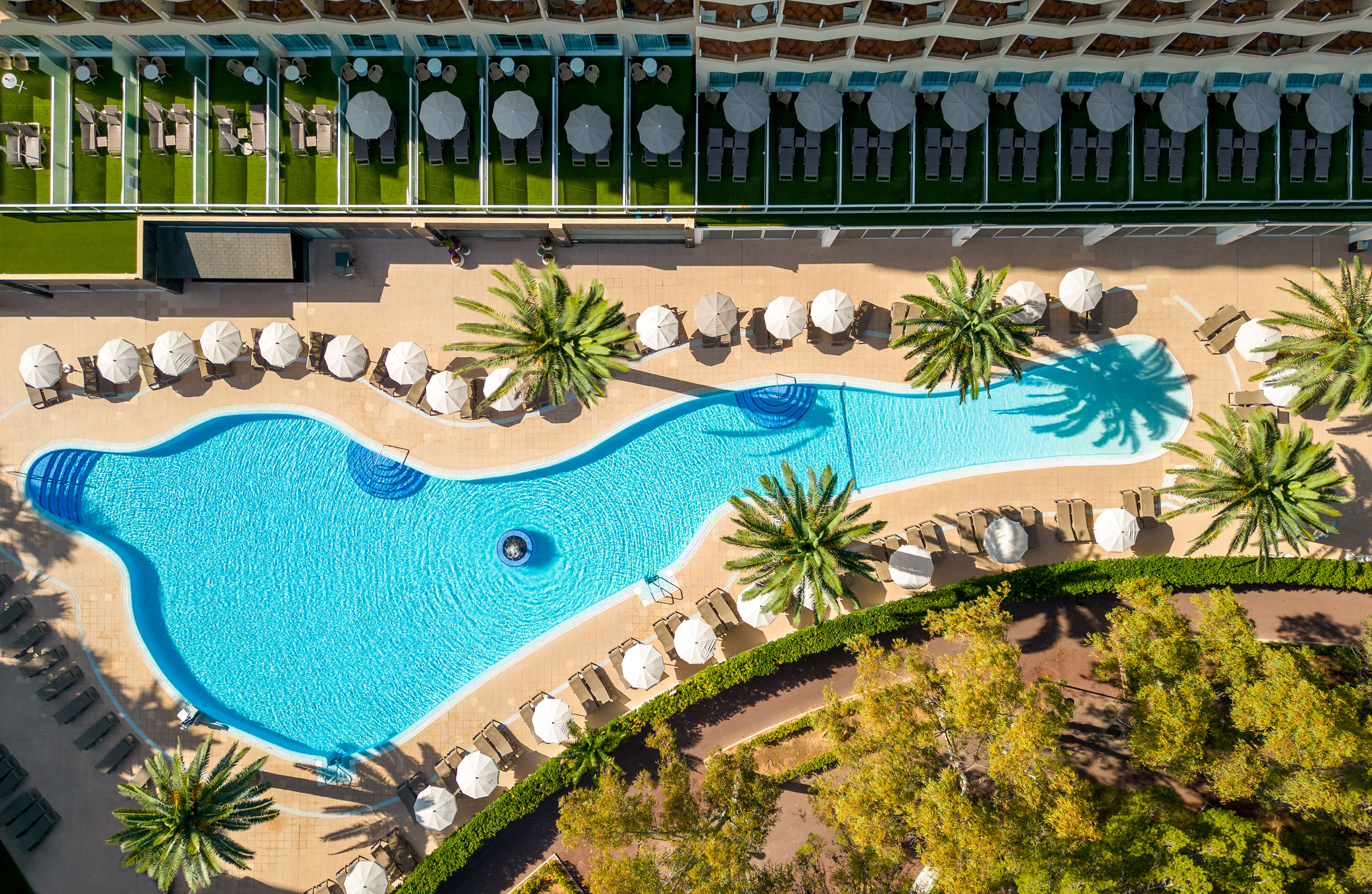 Aerial view of a resort swimming pool with palm trees, sun loungers, and umbrellas.