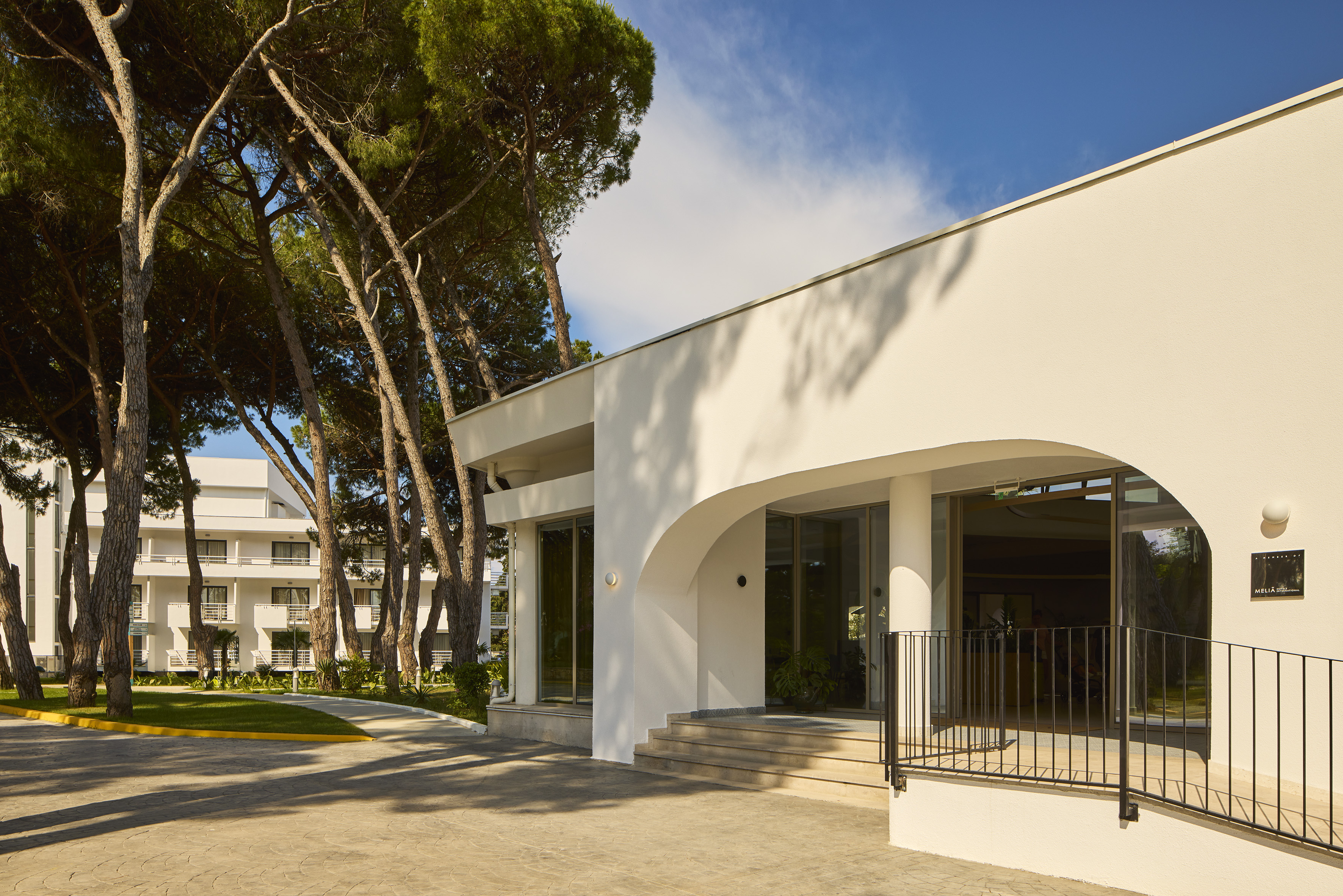 White modern hotel entrance with arched doorway and tall pine trees.