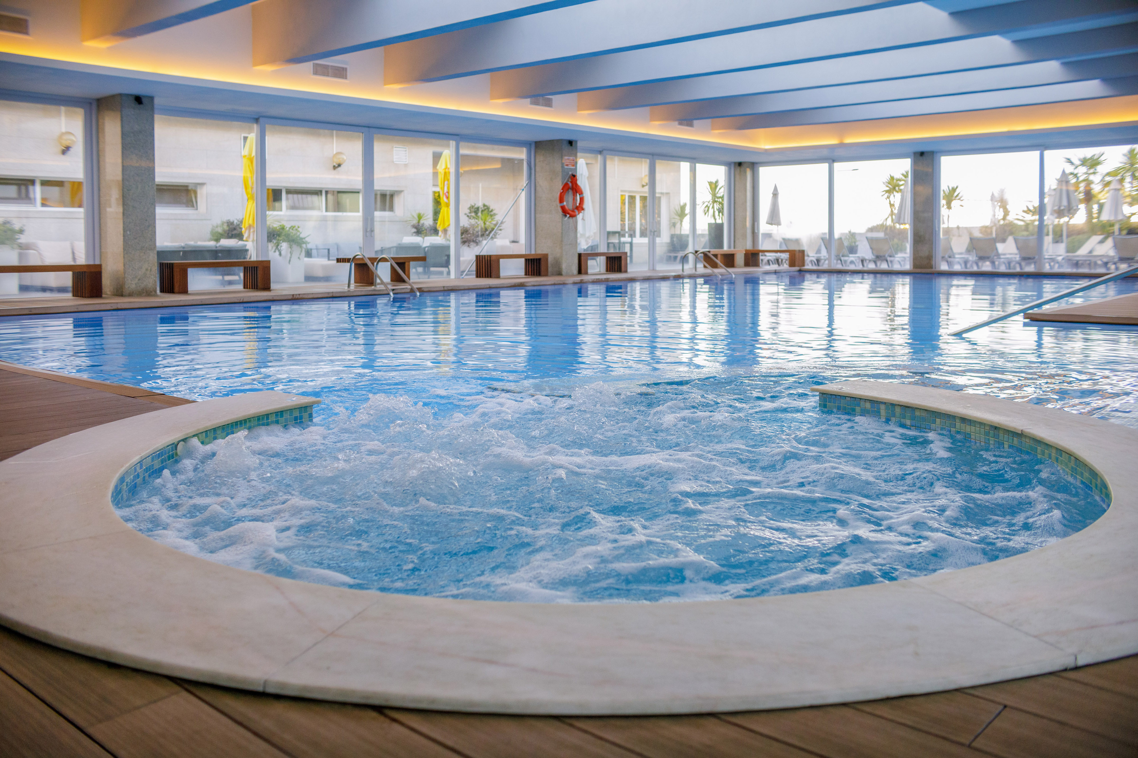 Hotel indoor swimming pool with hot tub, large windows, and wooden deck.