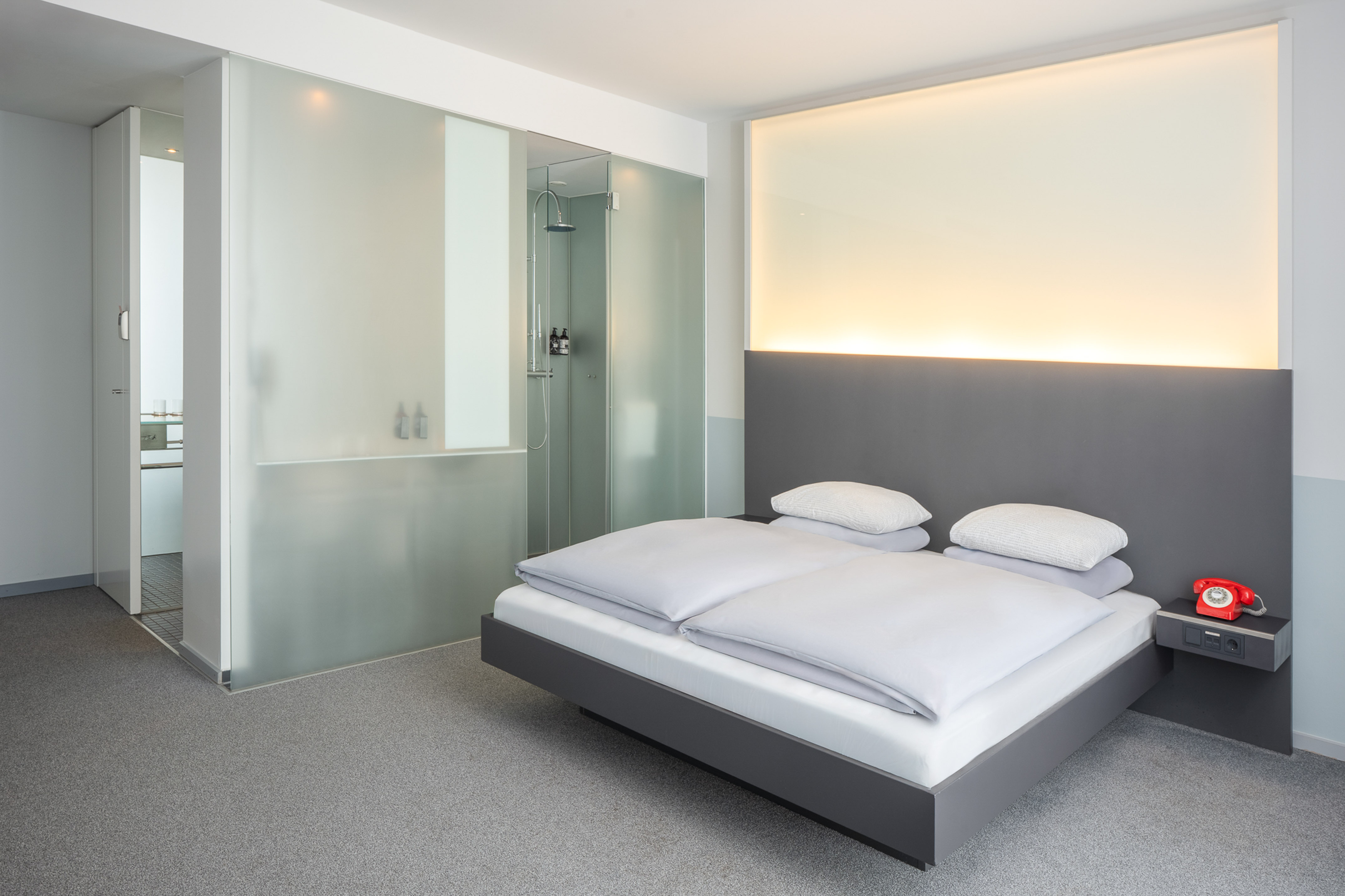 Minimalist hotel bedroom with gray bed, frosted glass shower, and red retro phone.