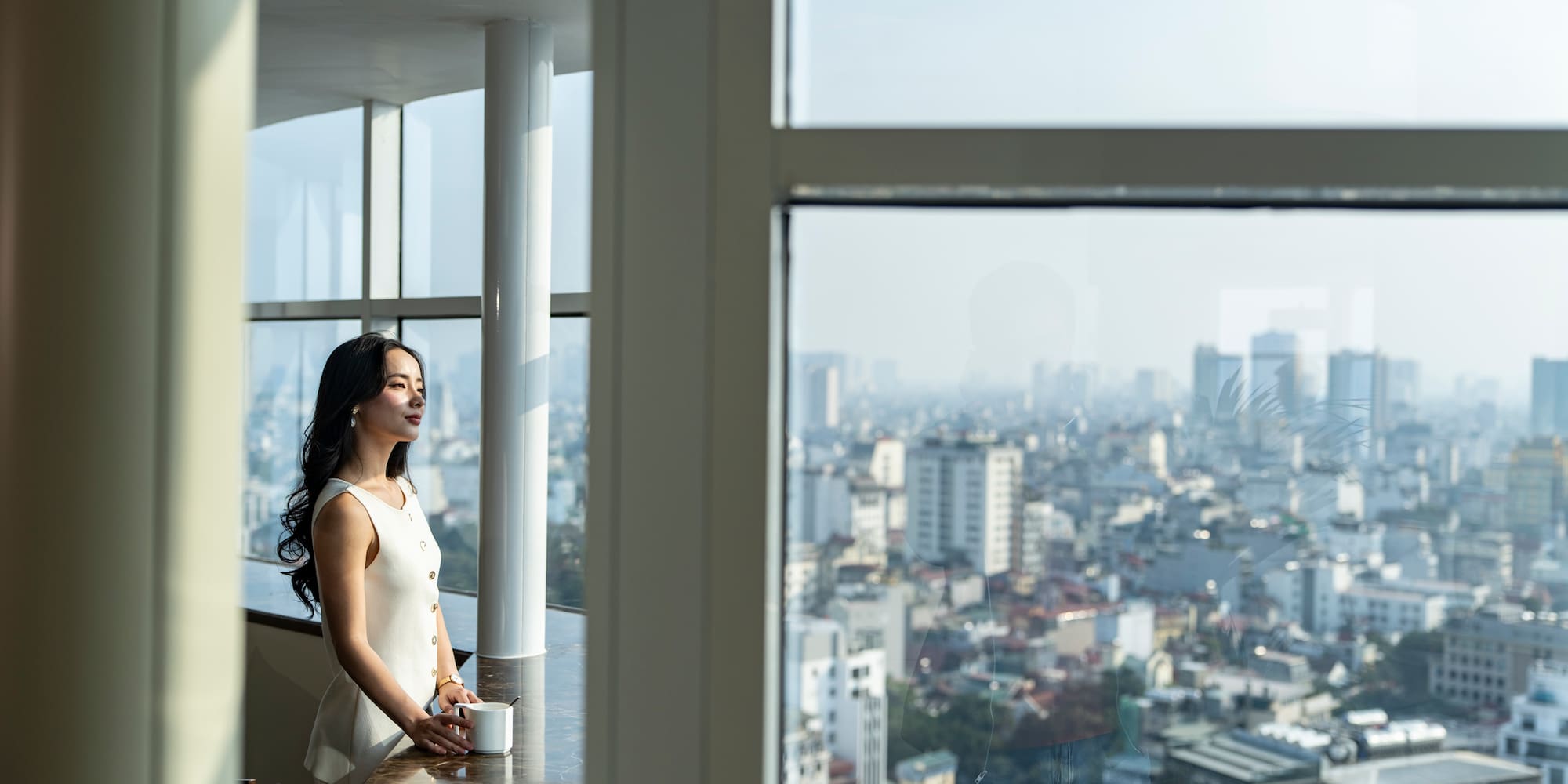 Woman with mug by window, overlooking a city skyline.