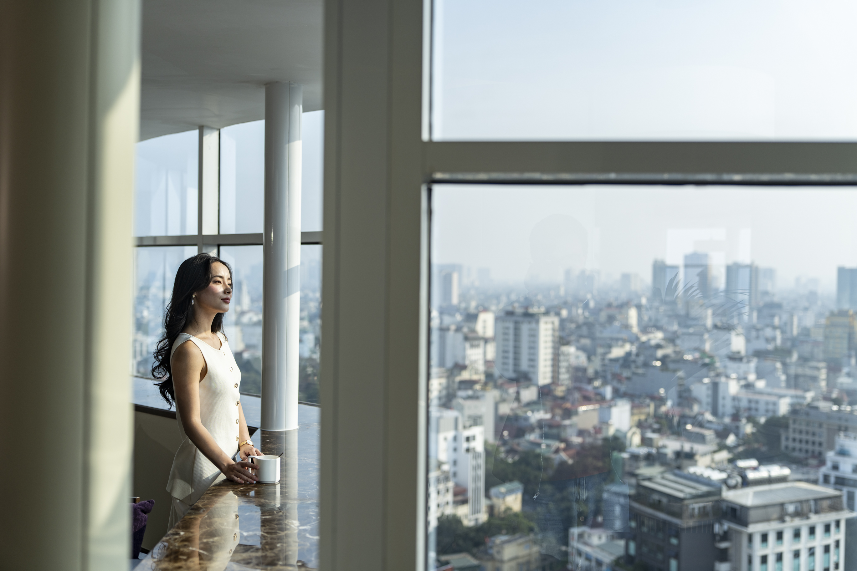 Woman with mug by window, overlooking a city skyline.