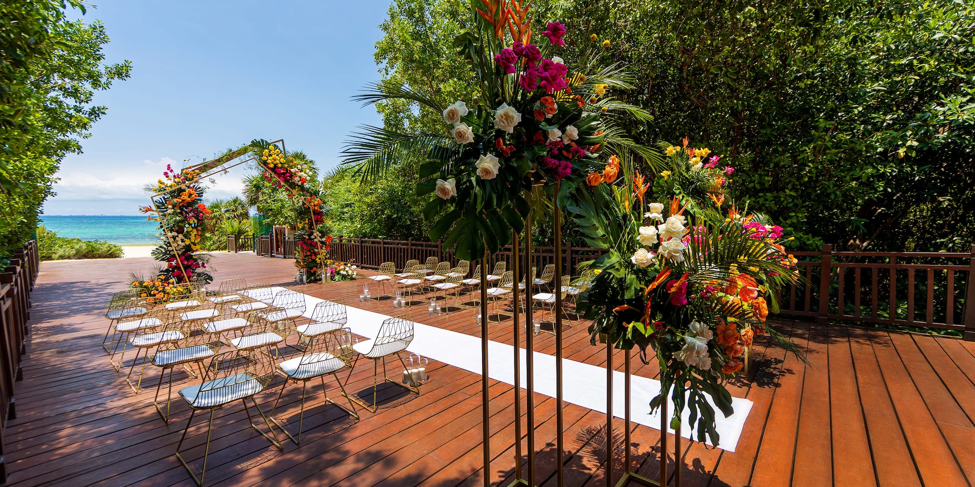 Beach wedding setup: wooden deck, floral arch, gold chairs, ocean backdrop.