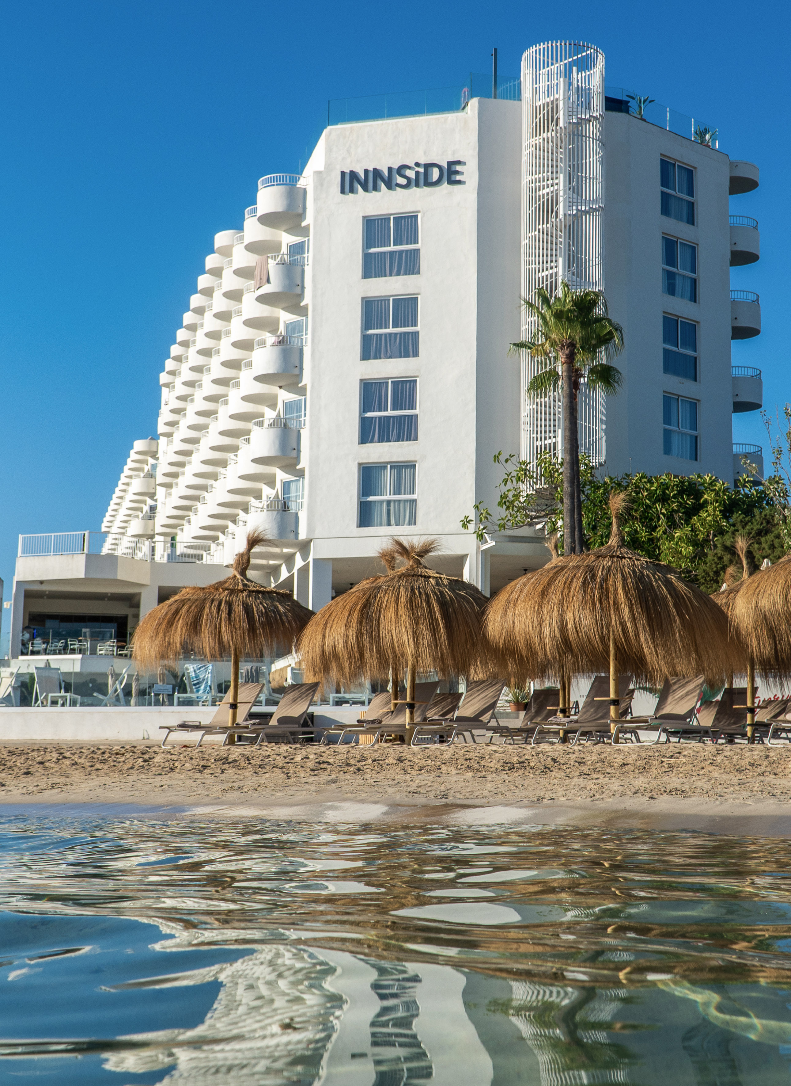 Waterfront INNSIDE hotel with beach umbrellas, lounge chairs, and clear blue sky.