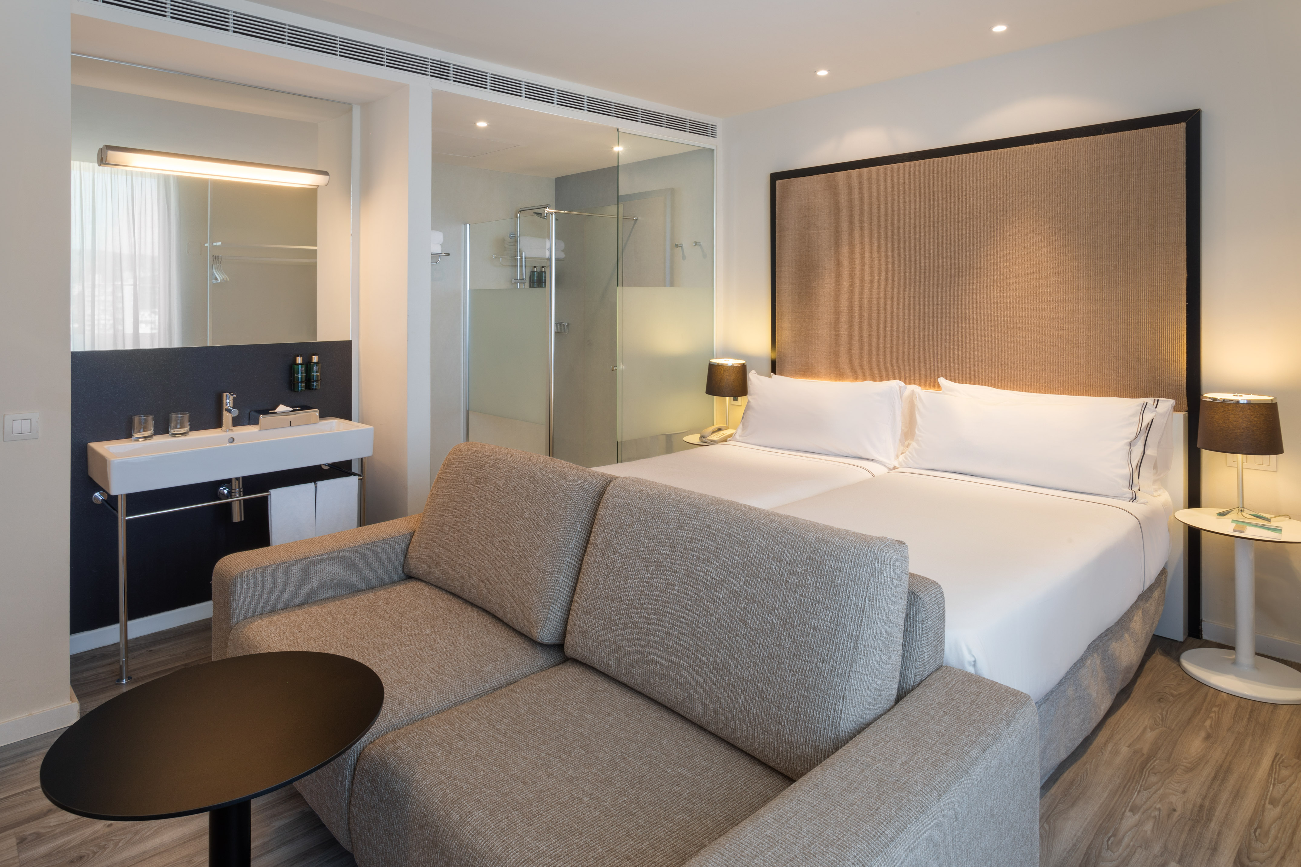 Contemporary hotel room with bed, sofa, and glass-walled bathroom.
