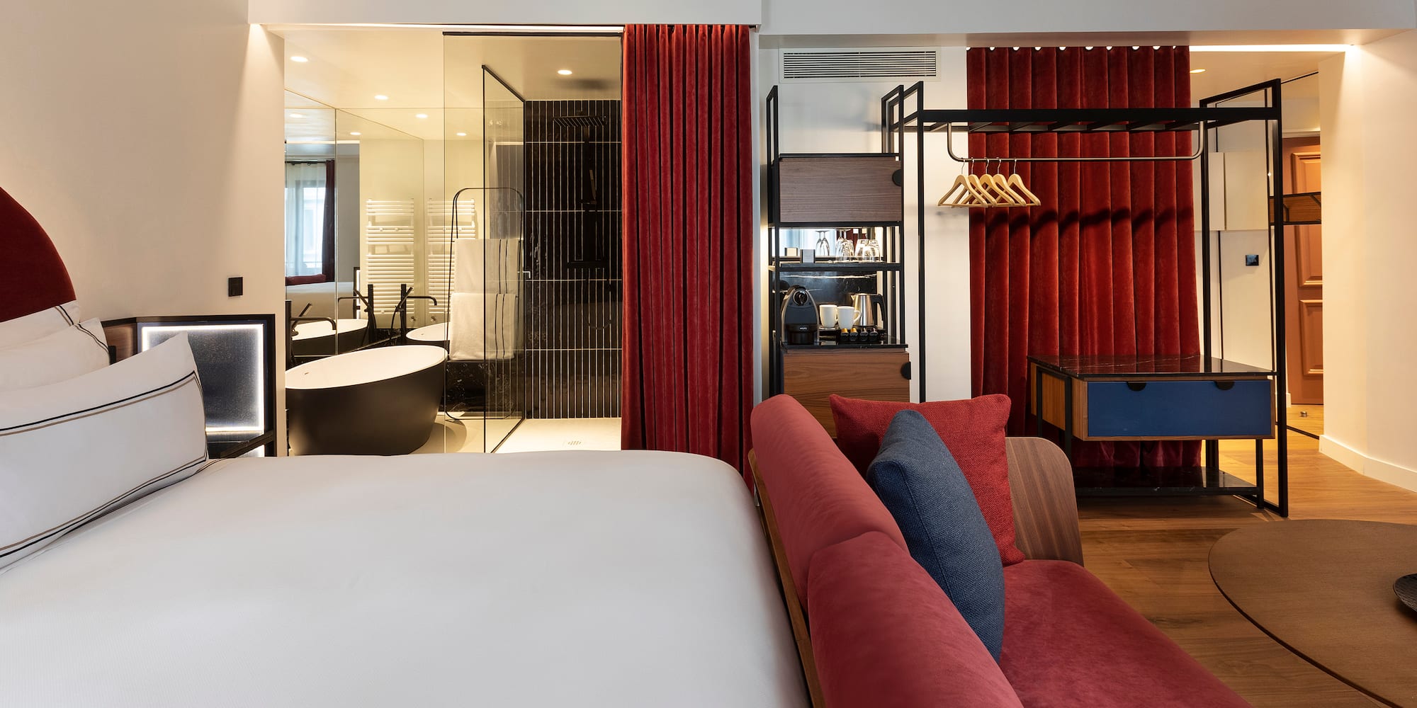Modern hotel suite with white bed, red sofa, open glass bathroom, and freestanding tub.