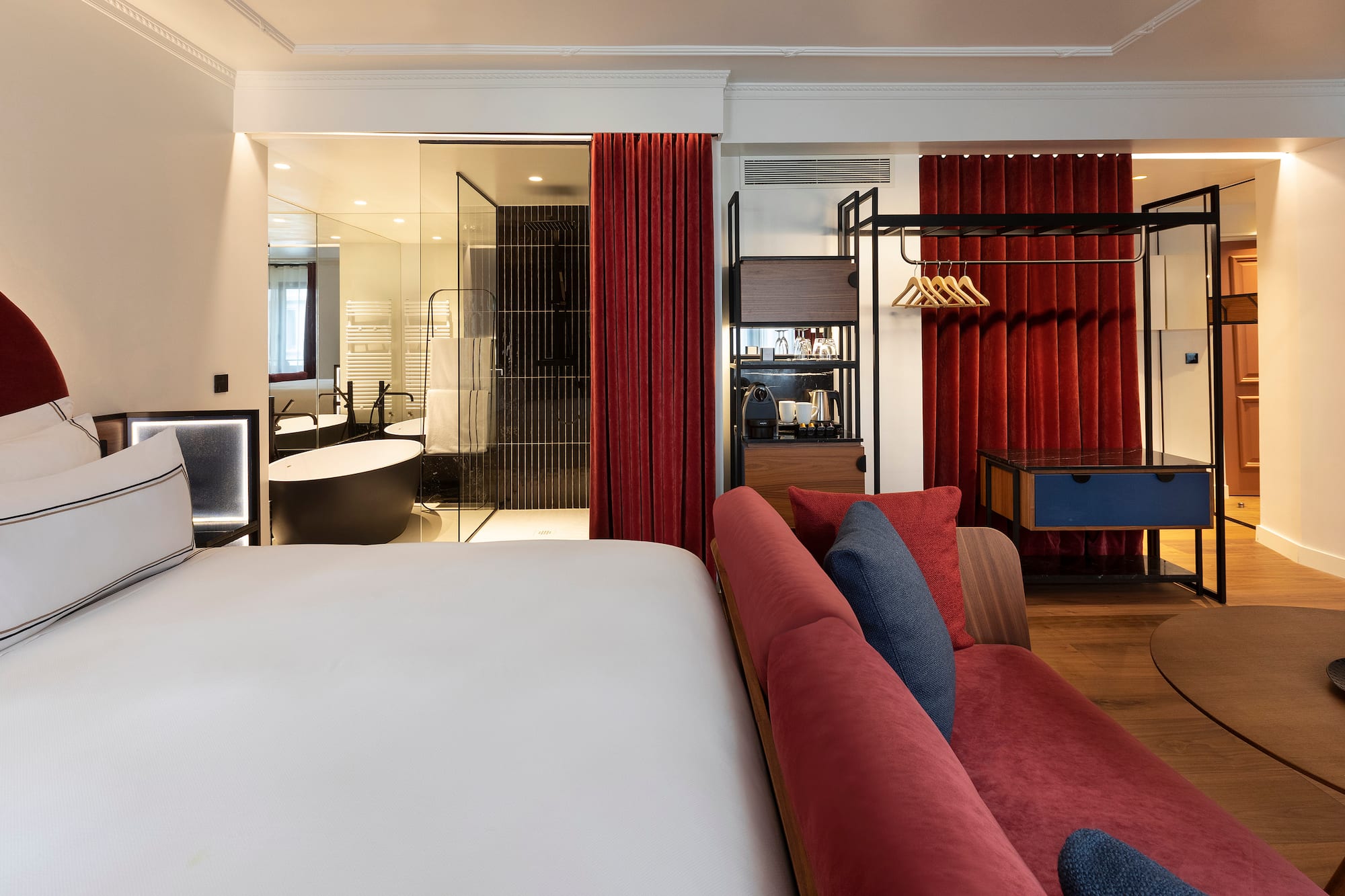 Modern hotel suite with white bed, red sofa, open glass bathroom, and freestanding tub.