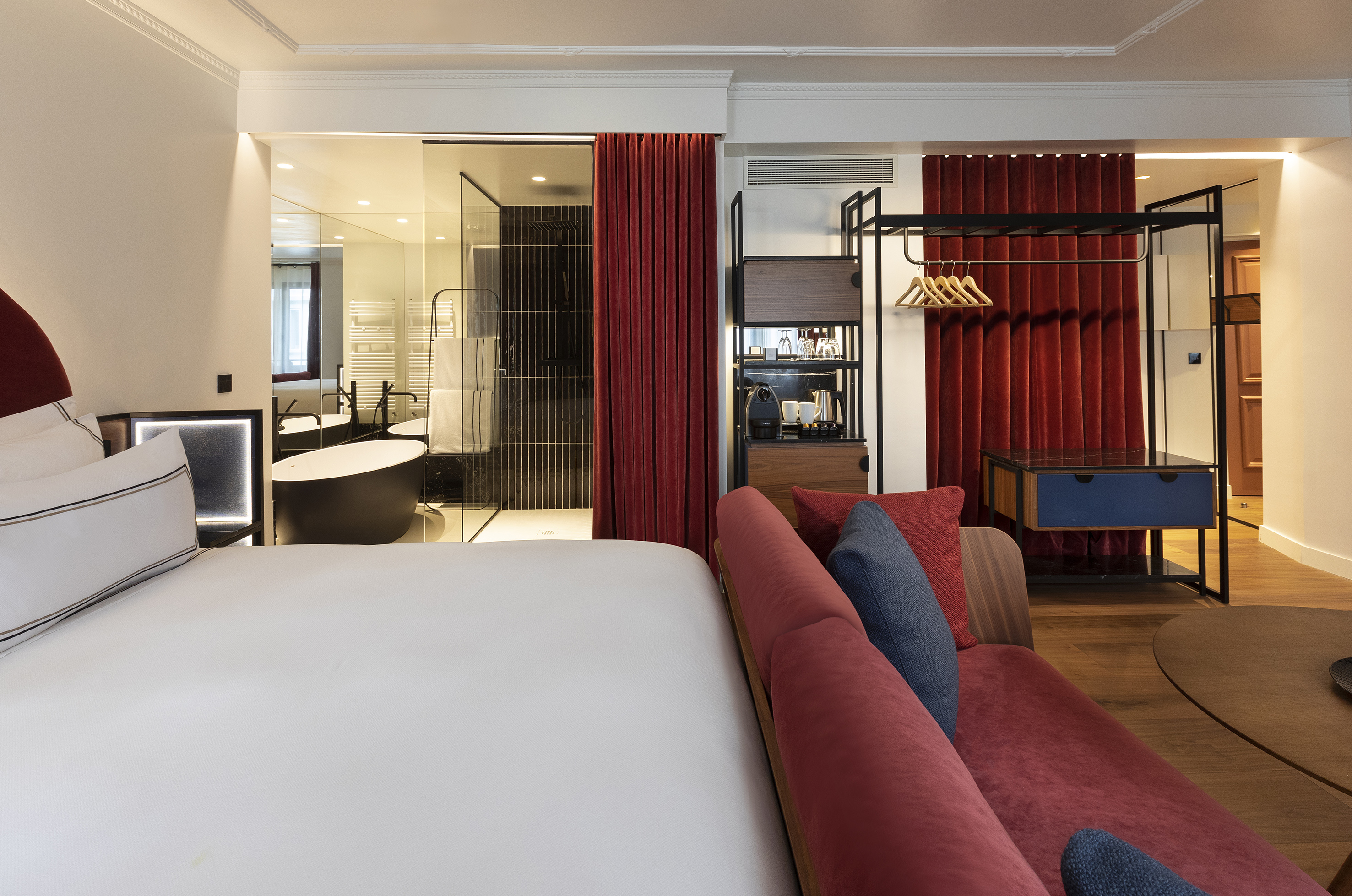 Modern hotel suite with white bed, red sofa, open glass bathroom, and freestanding tub.