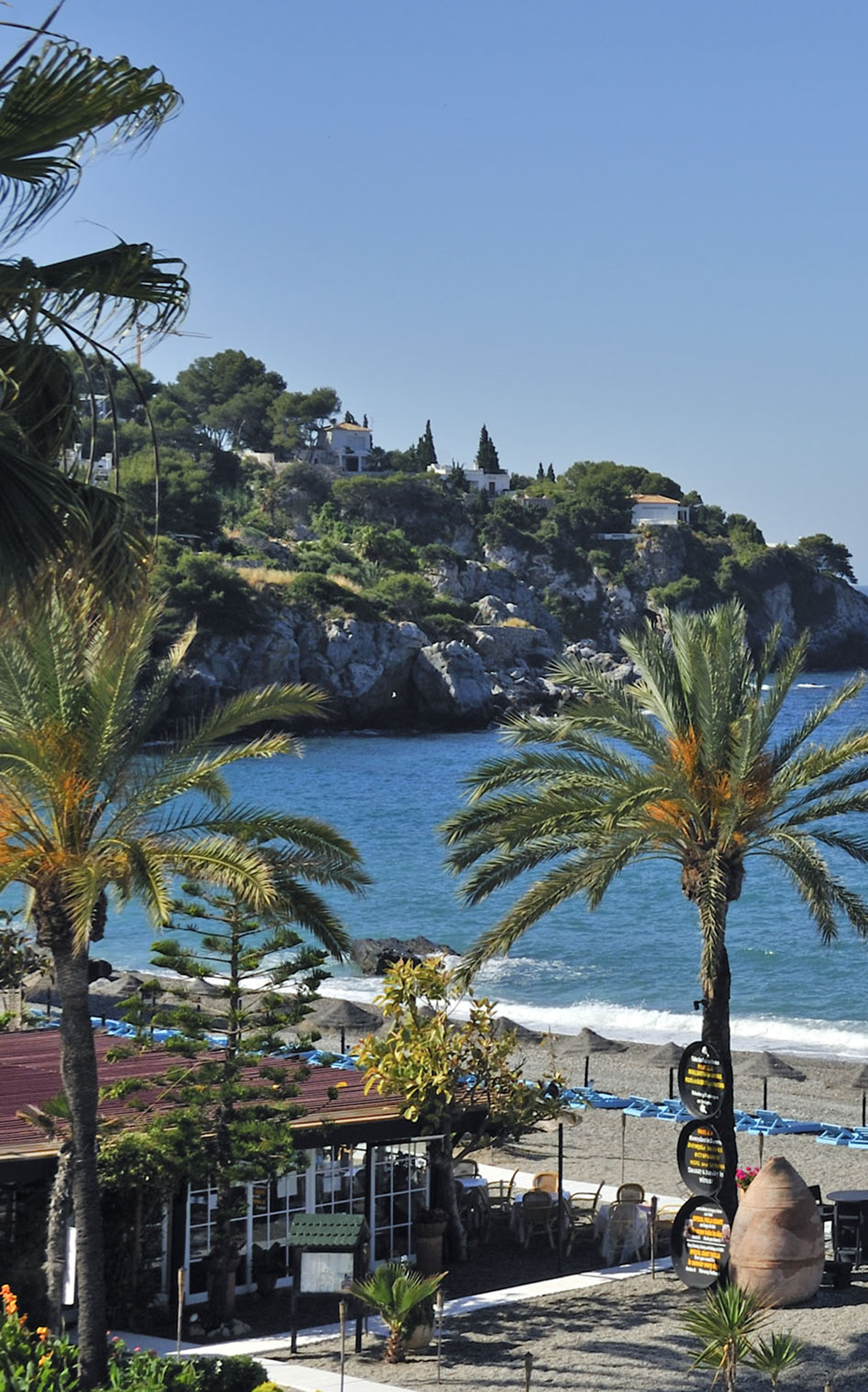 Palm-lined beach, blue ocean, rocky cliff, and seaside restaurant.