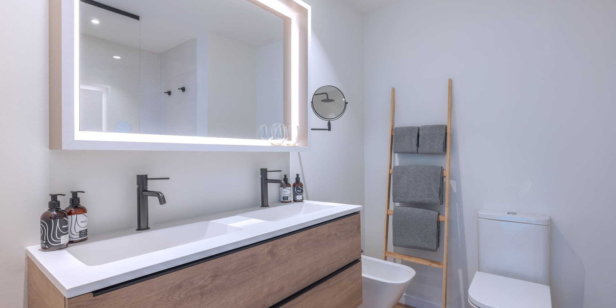 Baño moderno con mueble de madera, doble lavabo blanco y espejo iluminado.