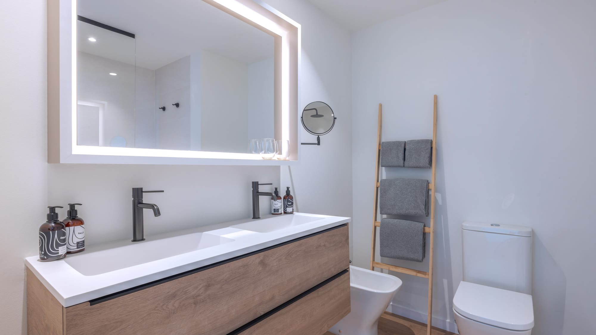 Baño moderno con mueble de madera, doble lavabo blanco y espejo iluminado.