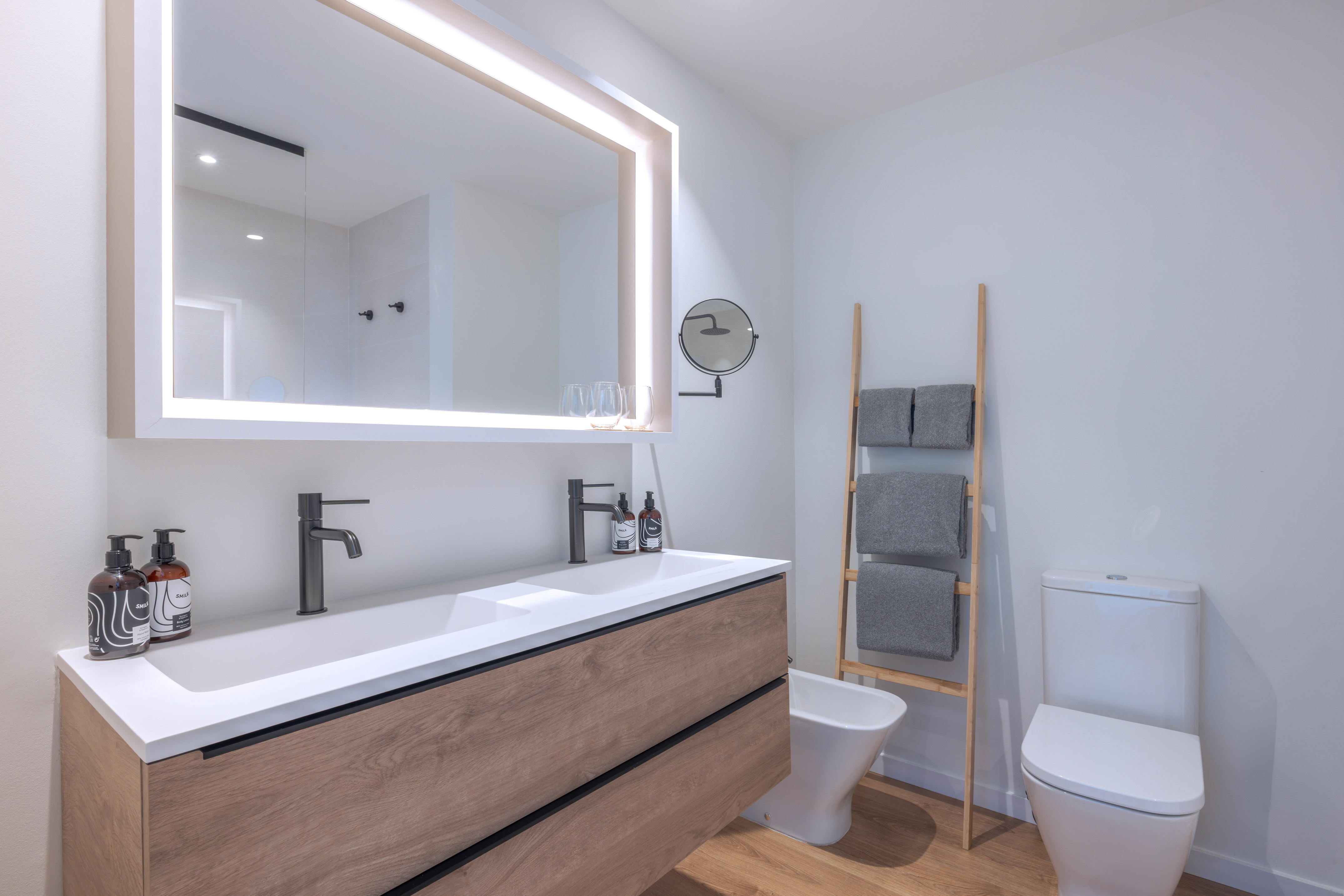 Baño moderno con mueble de madera, doble lavabo blanco y espejo iluminado.