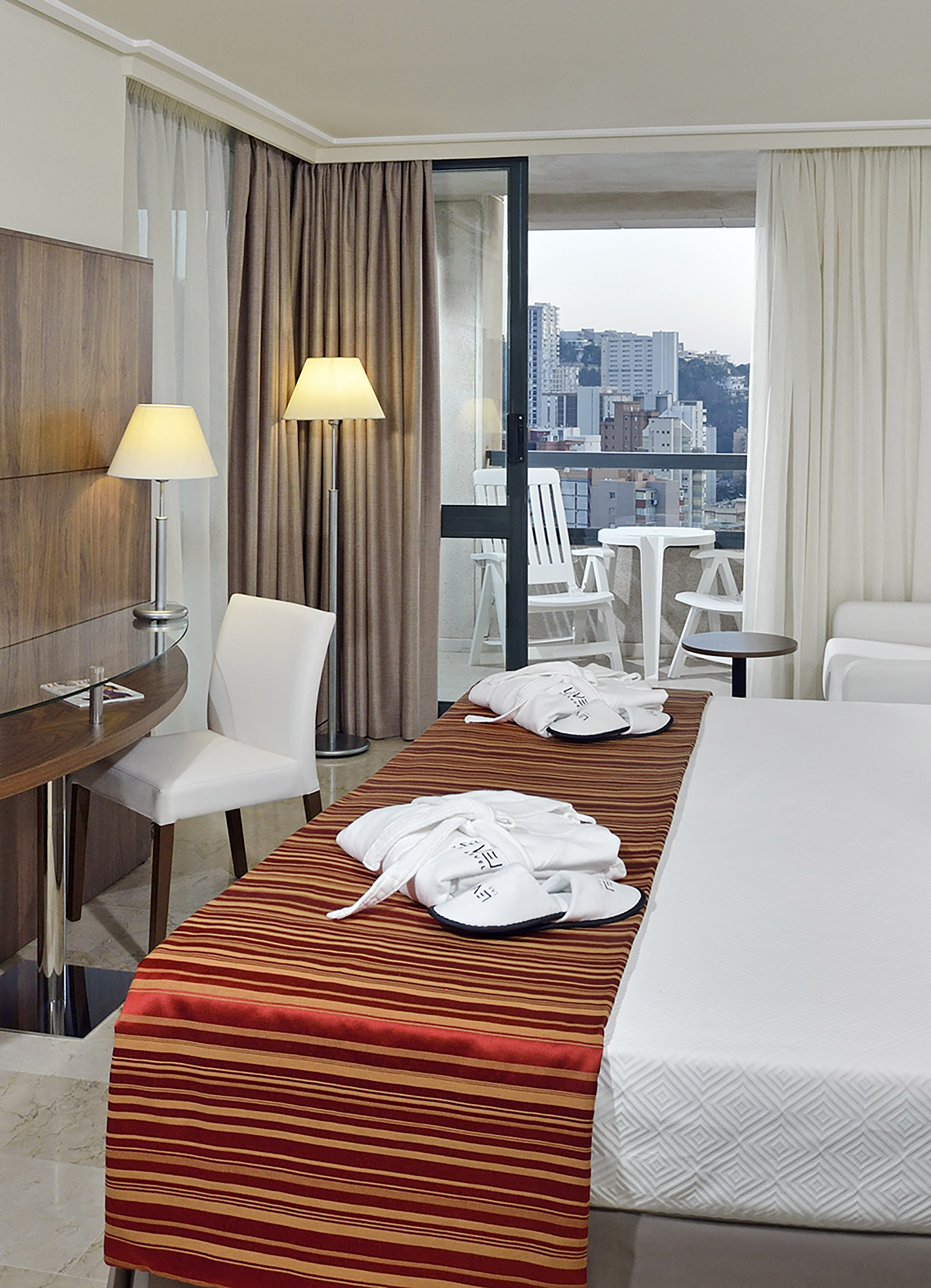 Modern hotel room with balcony, city view, bed with bathrobes and desk.