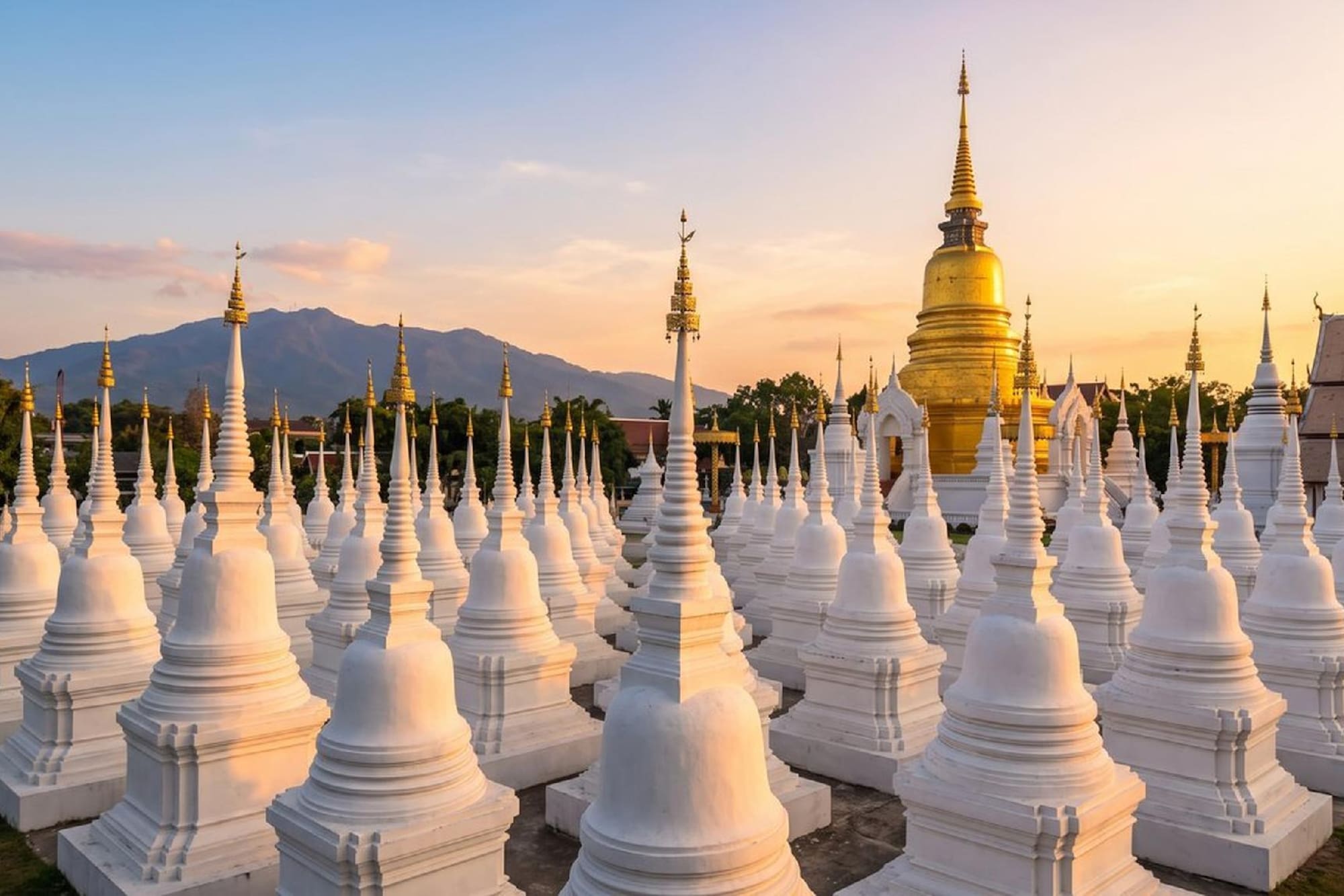 White stupas surround golden pagoda at a Thai temple with mountains, sunset.