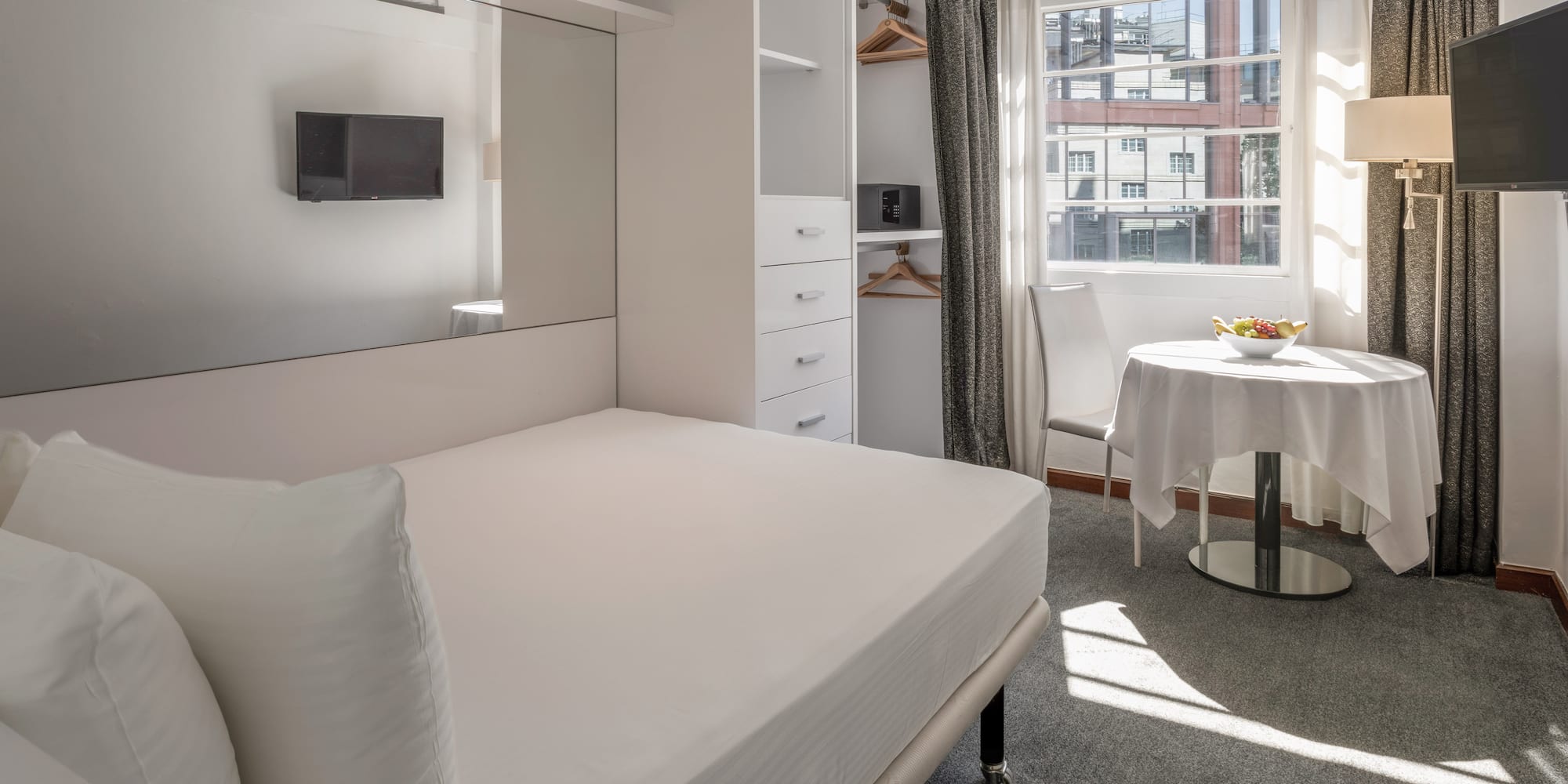Modern hotel studio with white bed, built-in storage, TV, and sunny window.