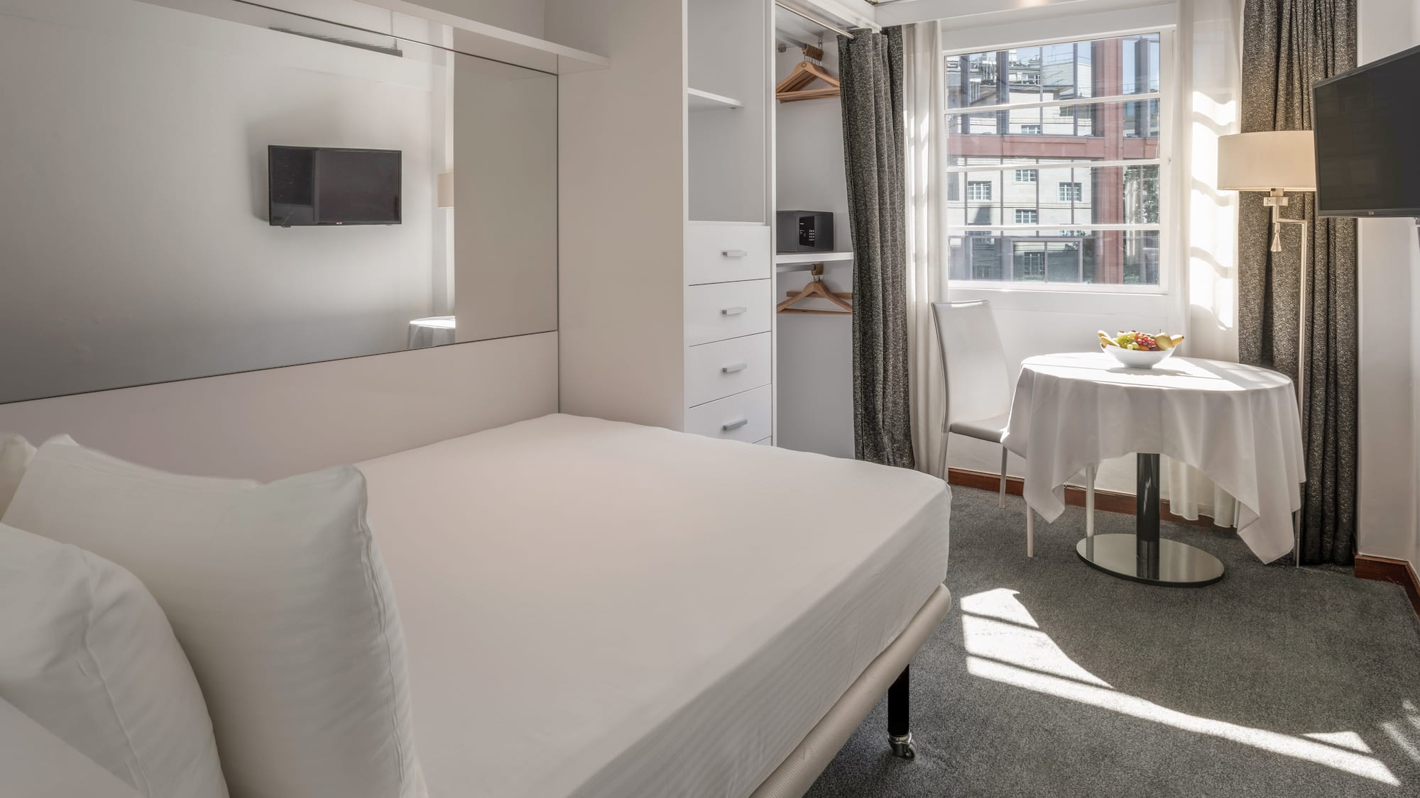 Modern hotel studio with white bed, built-in storage, TV, and sunny window.