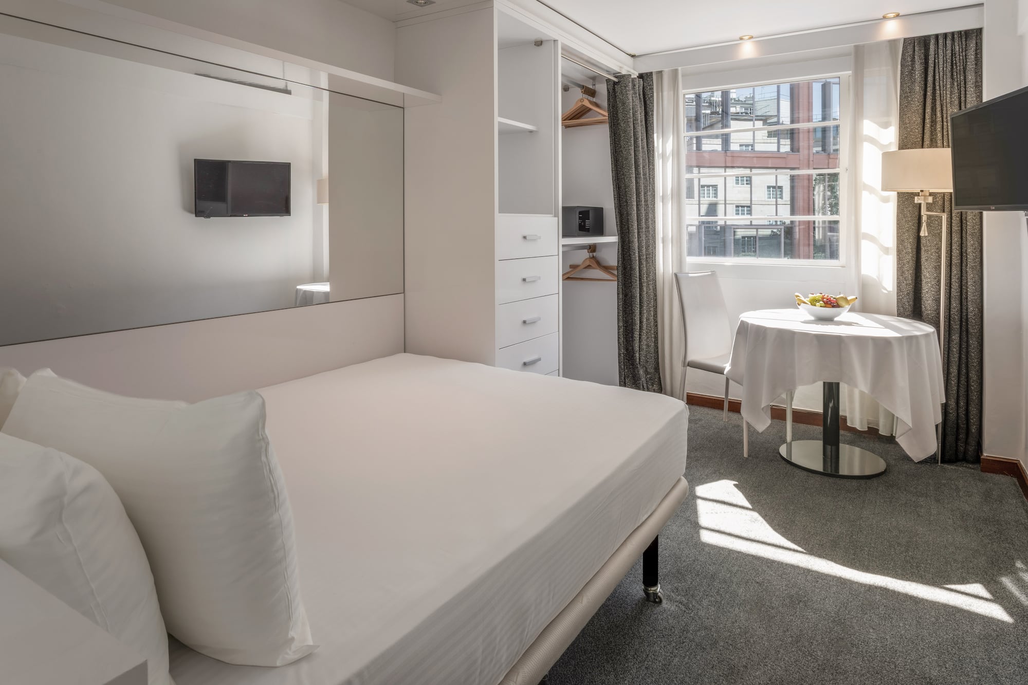 Modern hotel studio with white bed, built-in storage, TV, and sunny window.