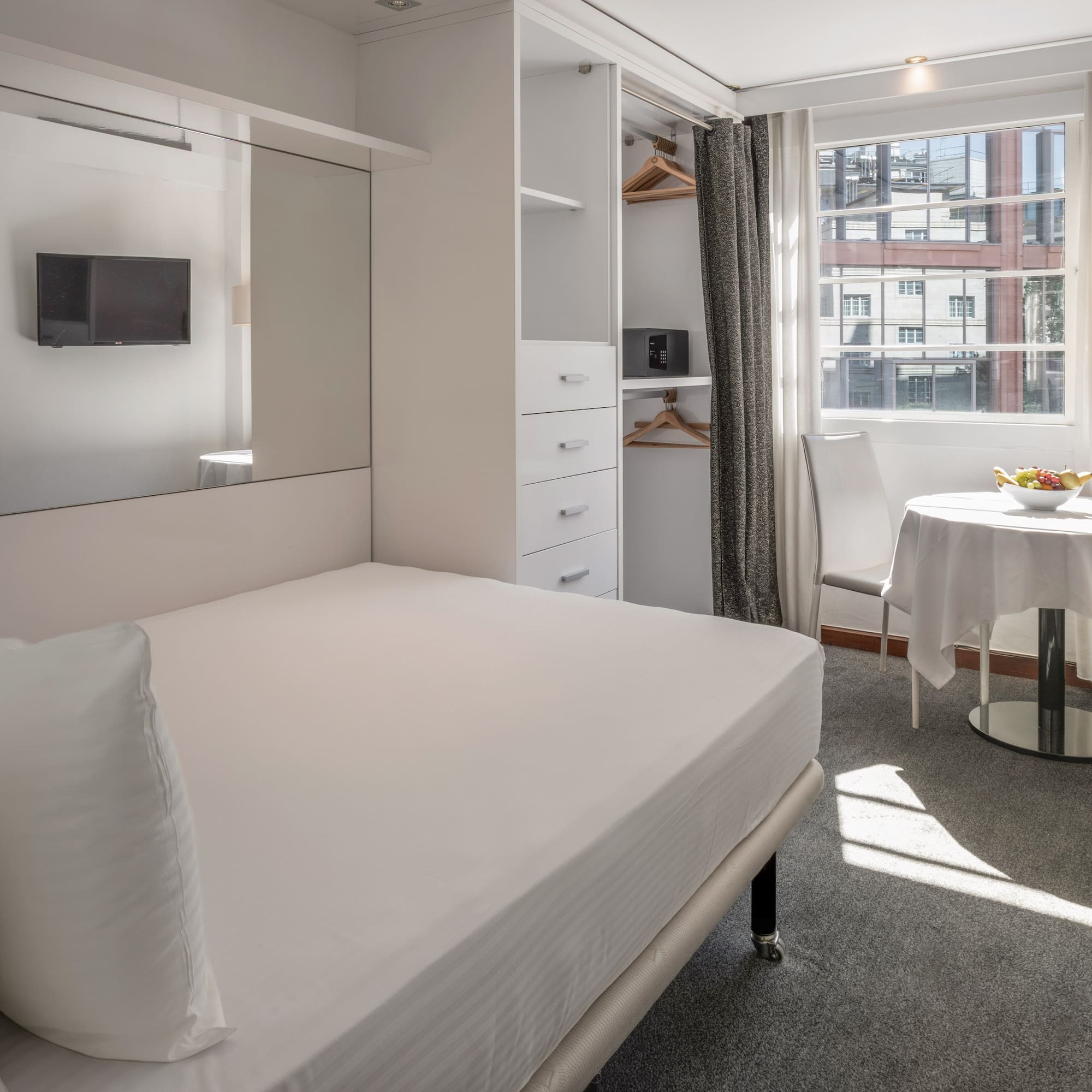 Modern hotel studio with white bed, built-in storage, TV, and sunny window.