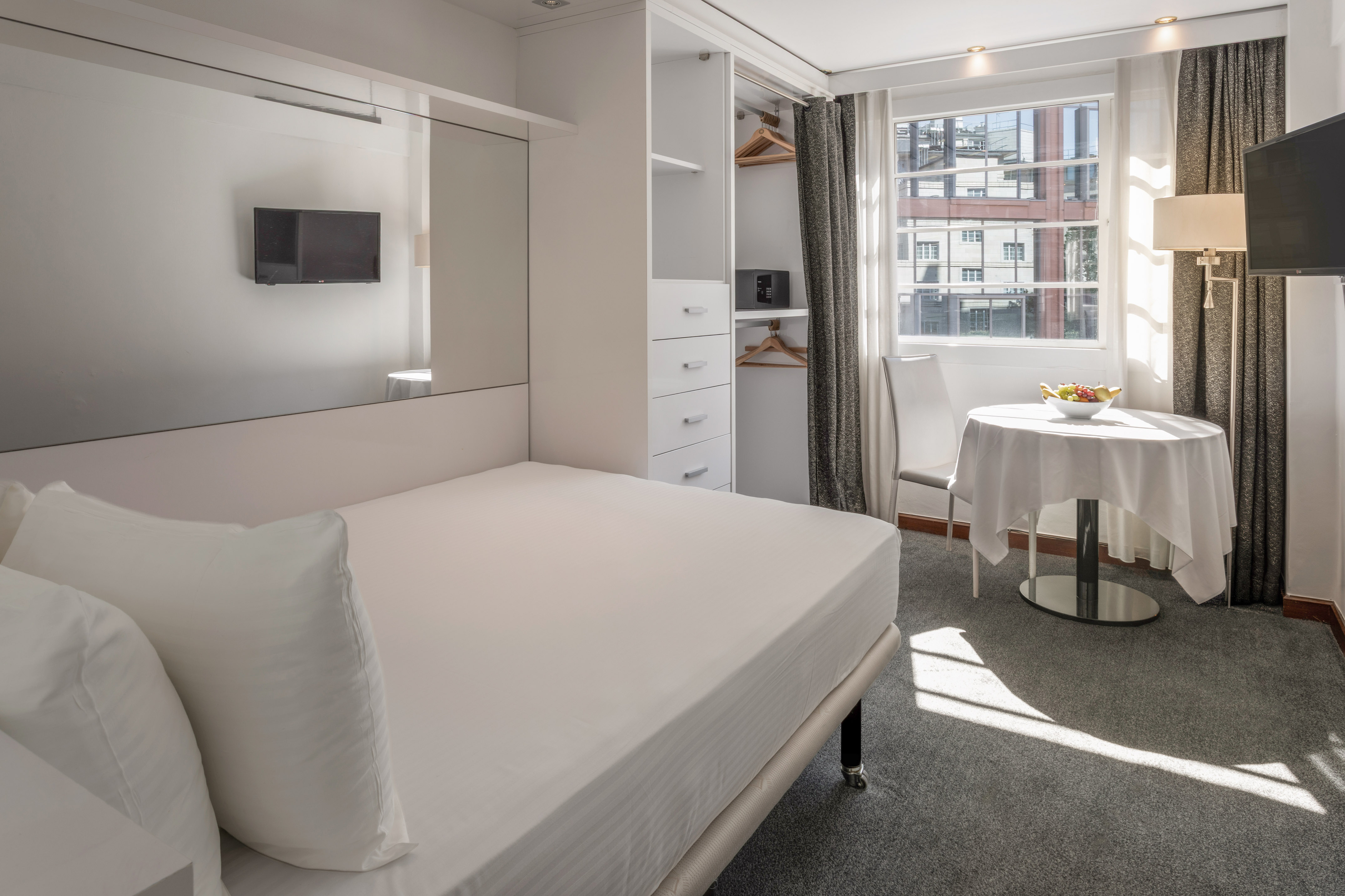 Modern hotel studio with white bed, built-in storage, TV, and sunny window.