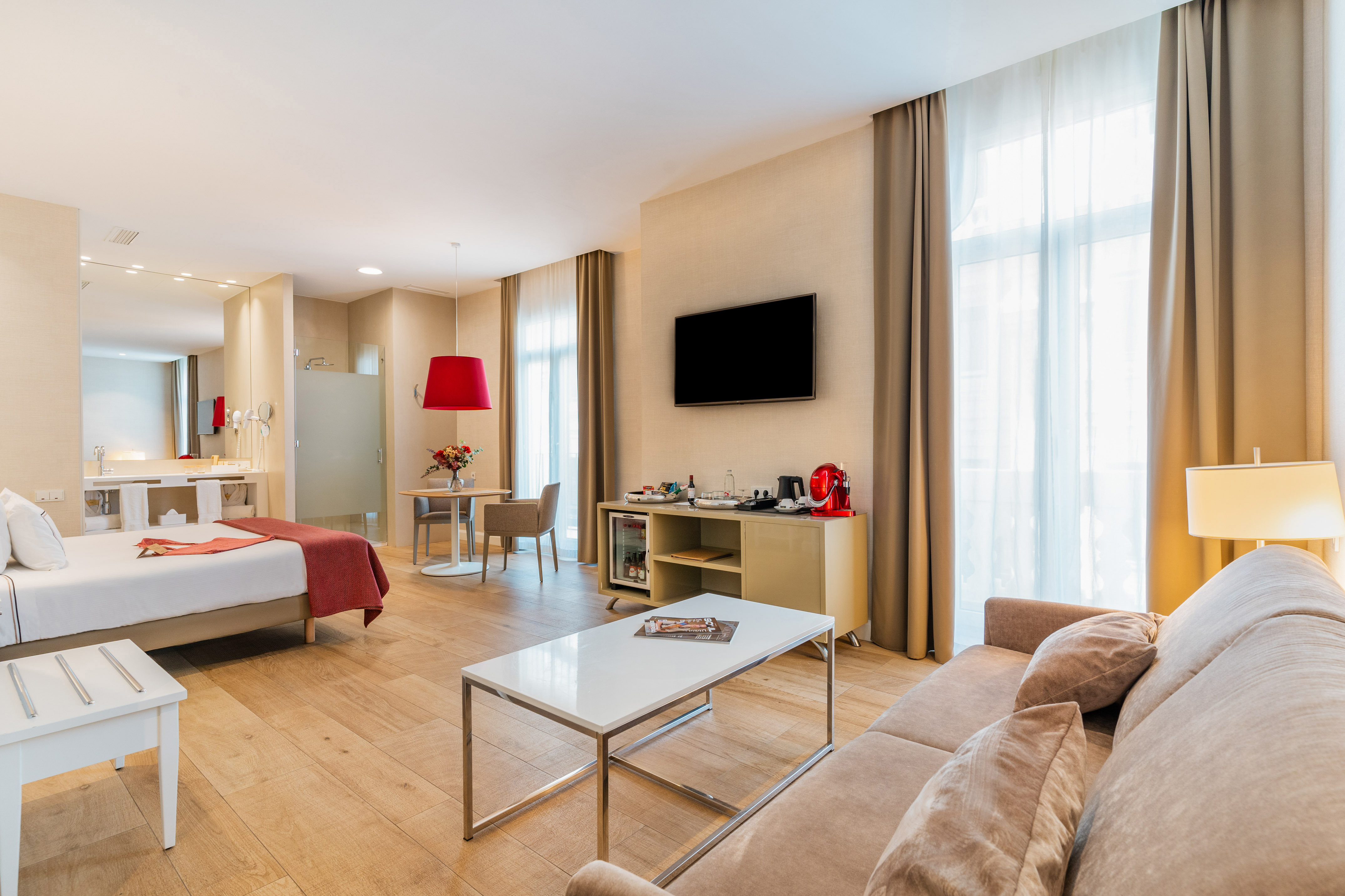 Spacious modern hotel suite with bed, sofa, TV, dining table, and minibar.