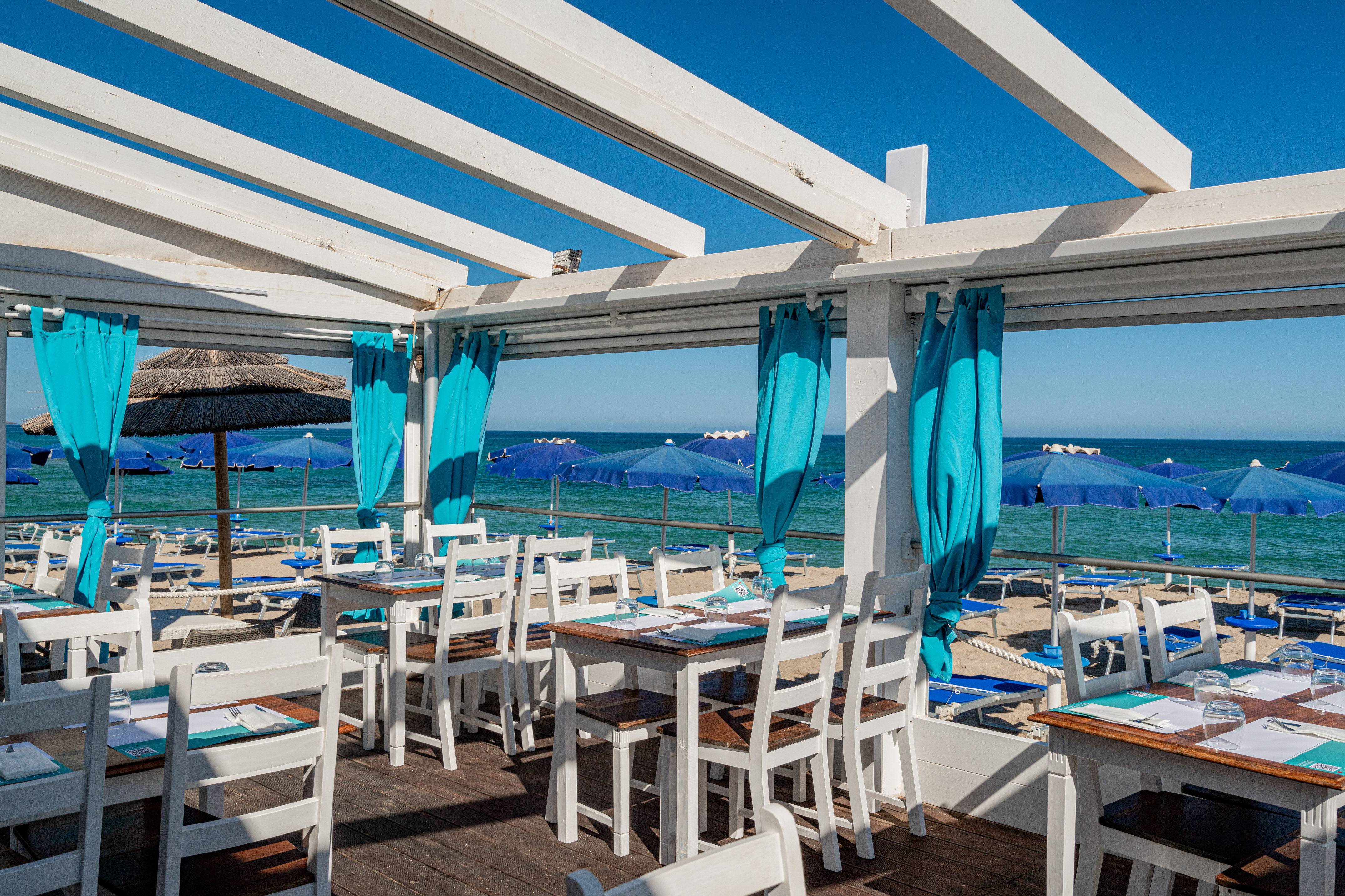Beachfront dining with ocean view, blue beach umbrellas, and white tables.