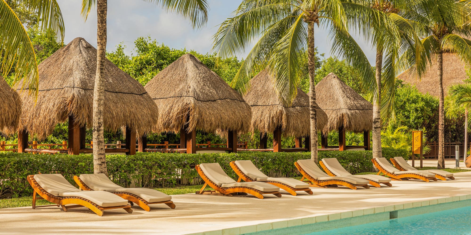 Tropical resort pool with lounge chairs, thatched huts, and palm trees.