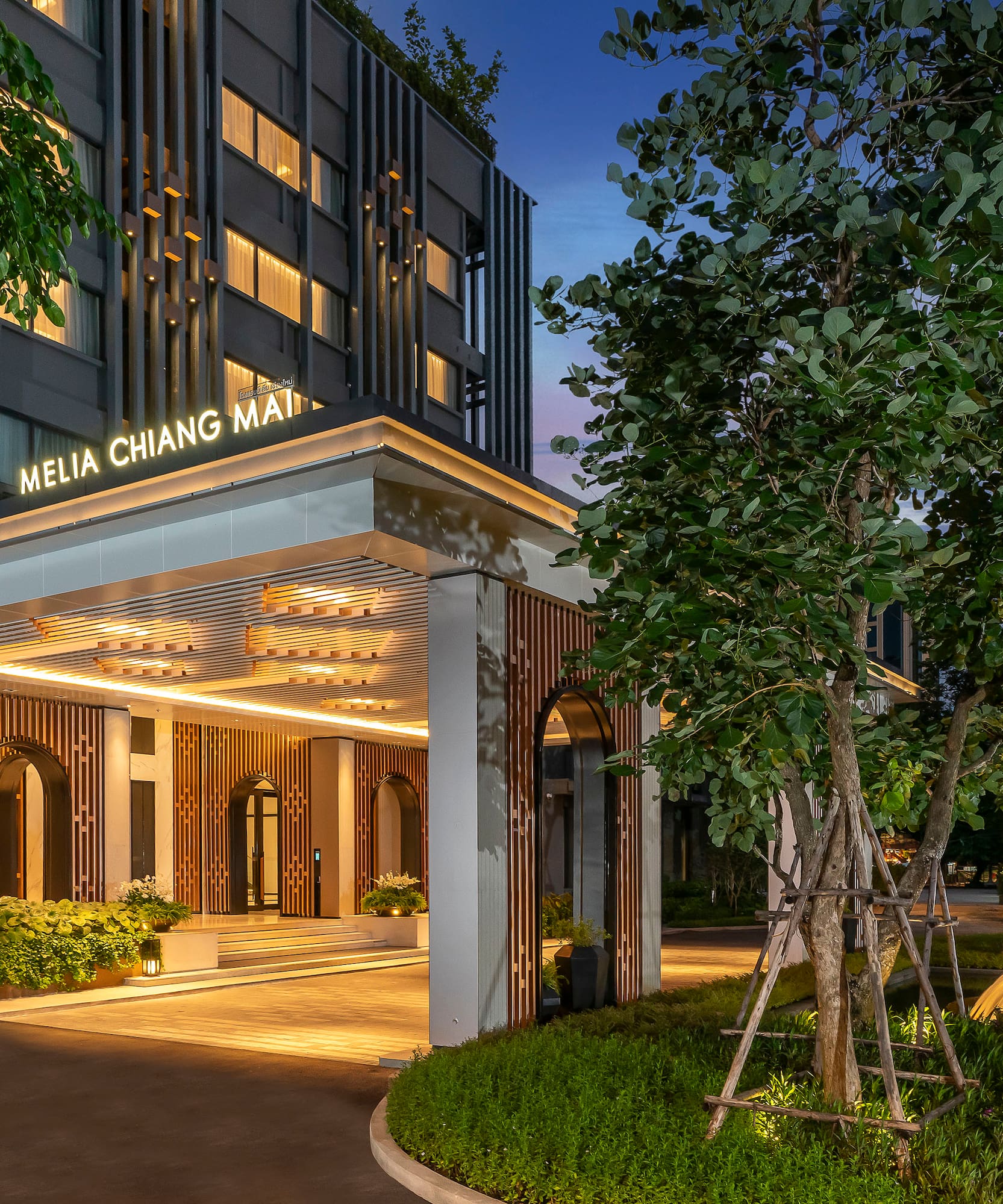 Illuminated Melia Chiang Mai hotel facade at twilight with lush trees and fountain.