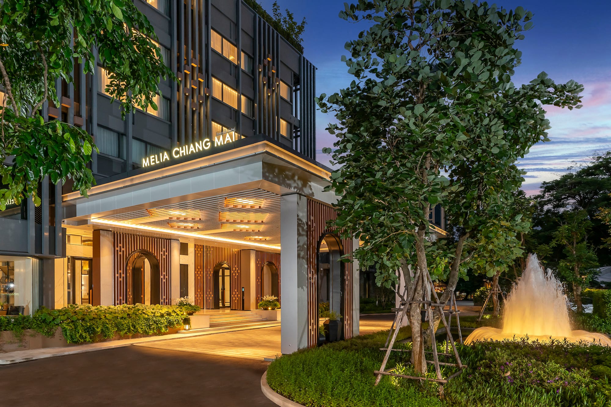 Illuminated Melia Chiang Mai hotel facade at twilight with lush trees and fountain.